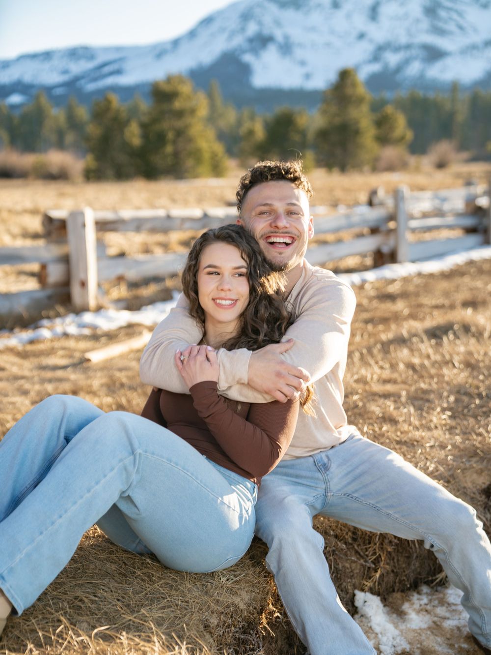 Couple sharing tender moments while sitting on ground with snow-capped mountains visible in distance.