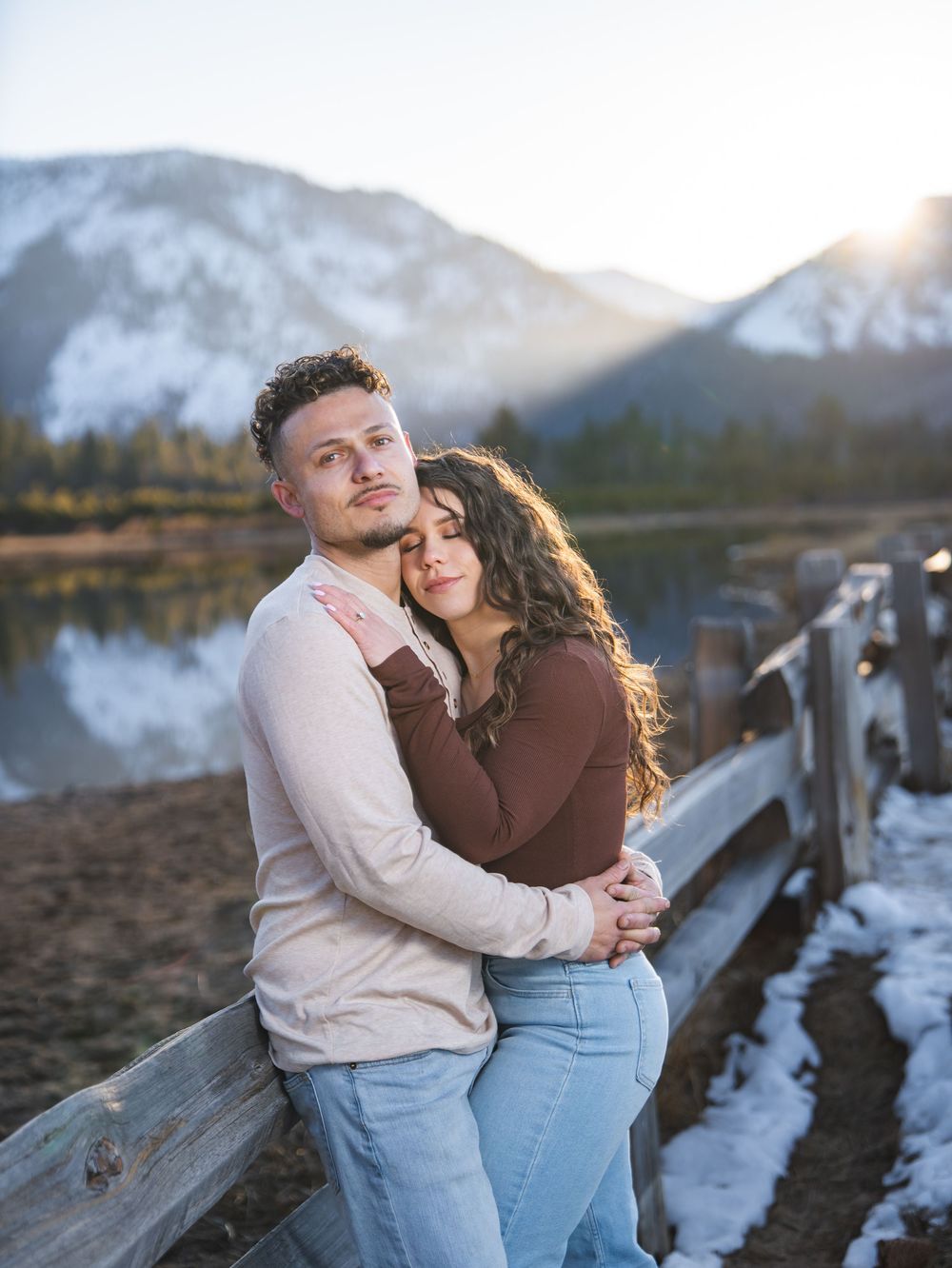 A romantic couple embraces by a mountain lake at sunset with snow-capped peaks in the background.