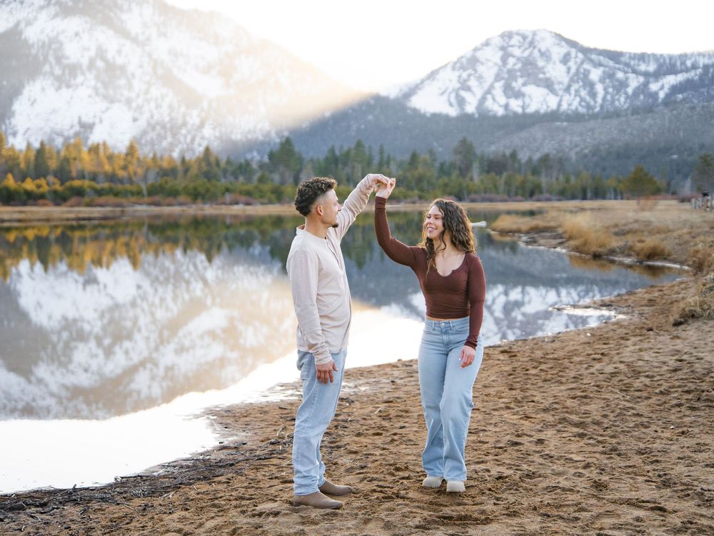 Two people walking hand in hand along a scenic mountain lake shoreline during golden hour.