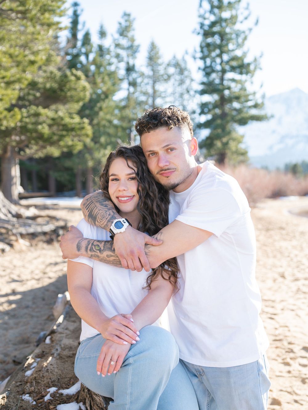 A couple in white shirts embrace on a sandy beach with pine trees in the background during golden hour.