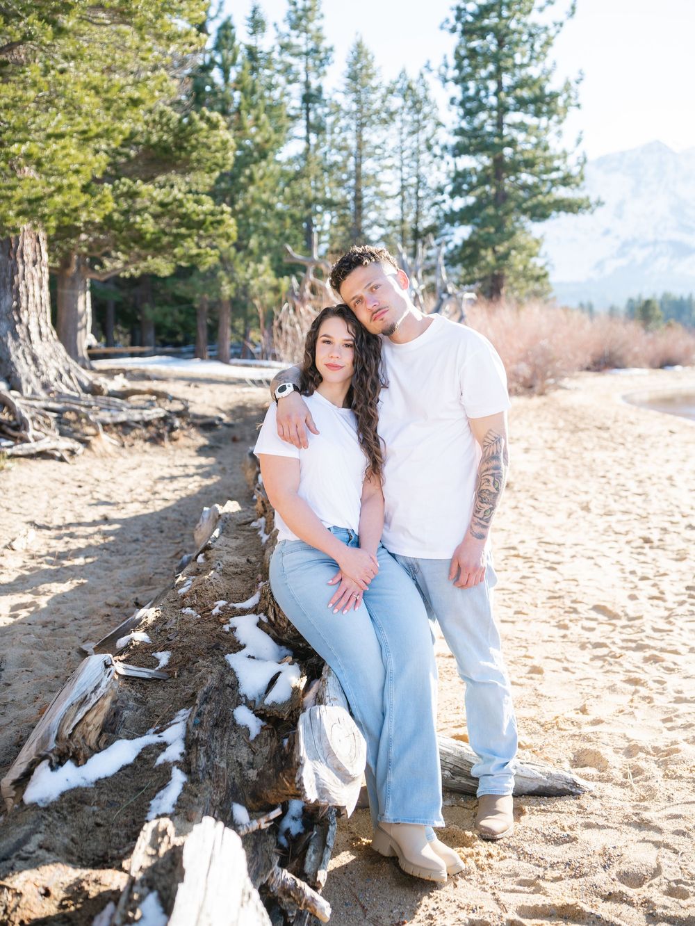 A couple poses together on a sandy beach with pine trees and mountains in the background.