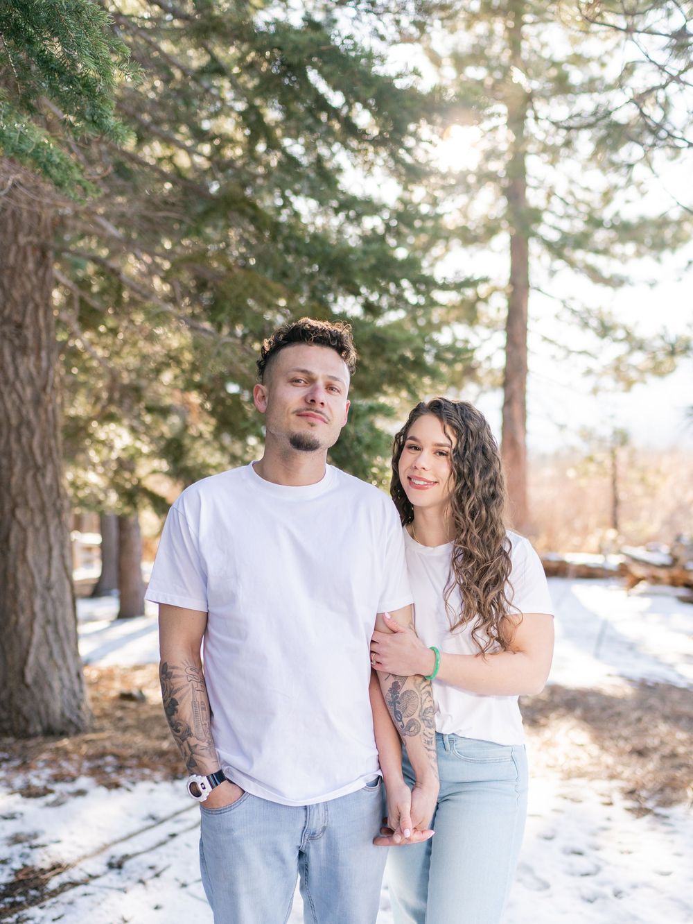 A couple in matching white tops and light denim poses together in a snowy forest setting with sunlit trees.