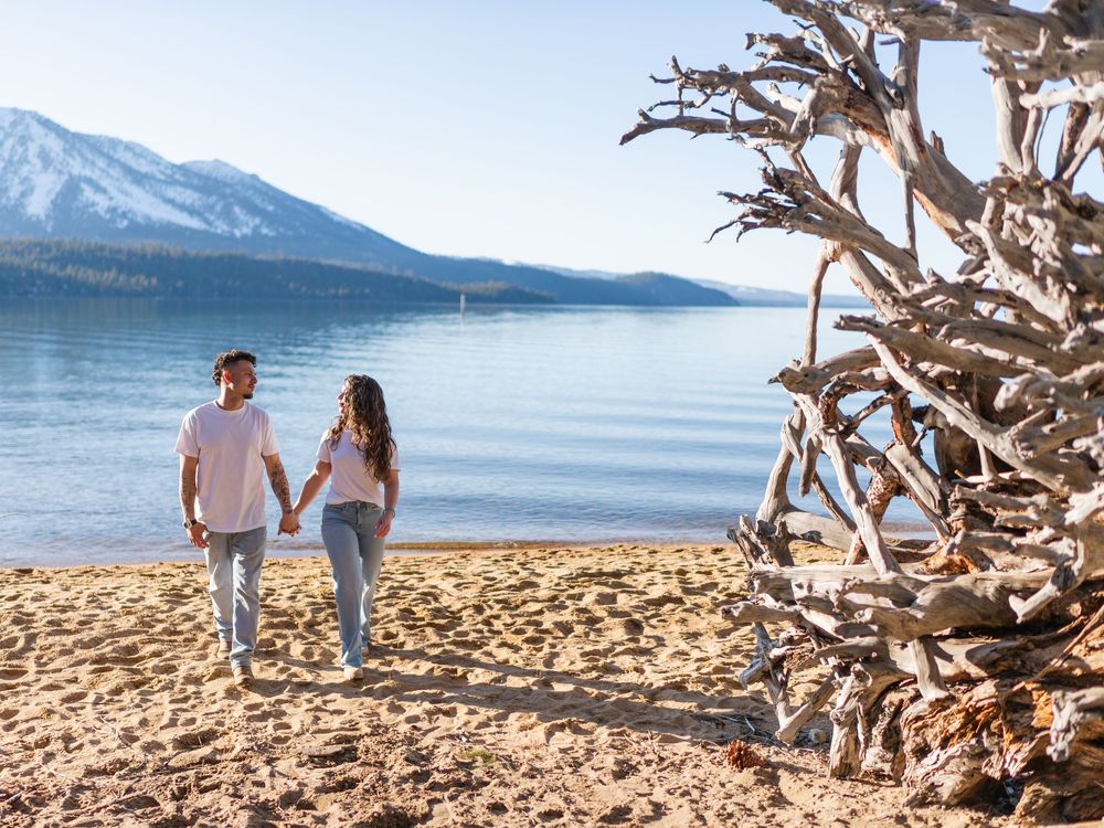 A couple walks hand in hand along a scenic beach with mountains and lake in the background.
