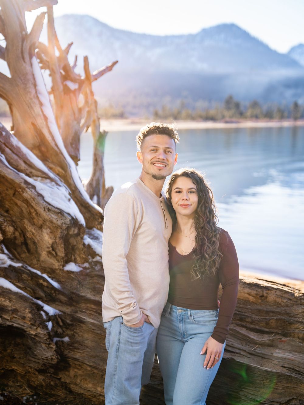 A couple poses together by a large driftwood log on a serene lake shore with mountains in the background.