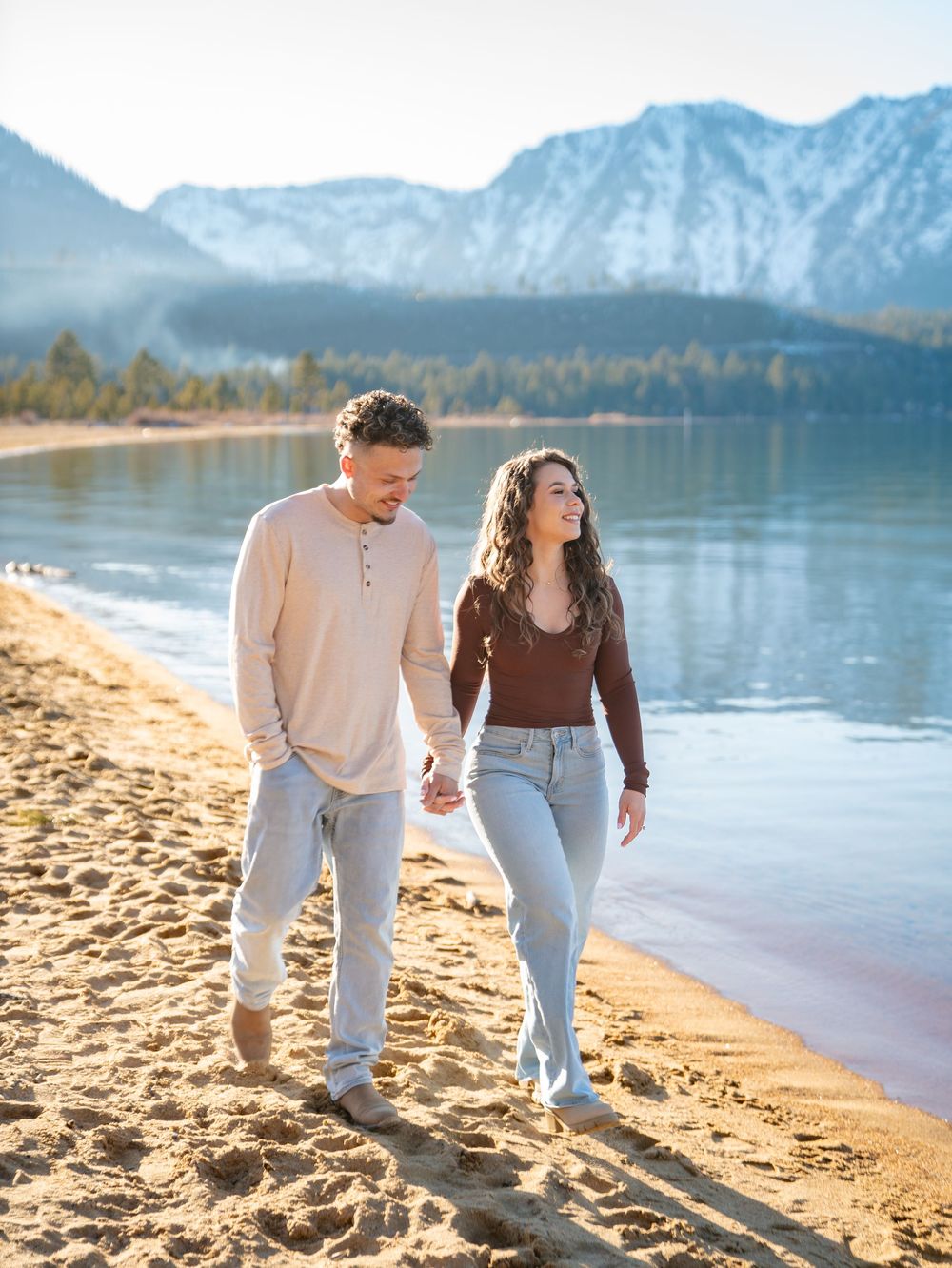 Two people walking hand in hand along a sandy beach with mountains and lake in the background during sunset.