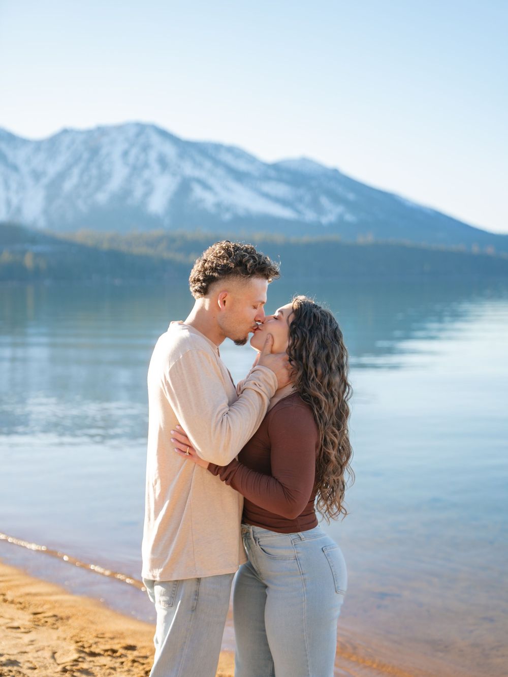 A couple shares a romantic moment on a sandy beach with snow-capped mountains and a serene lake in the background.