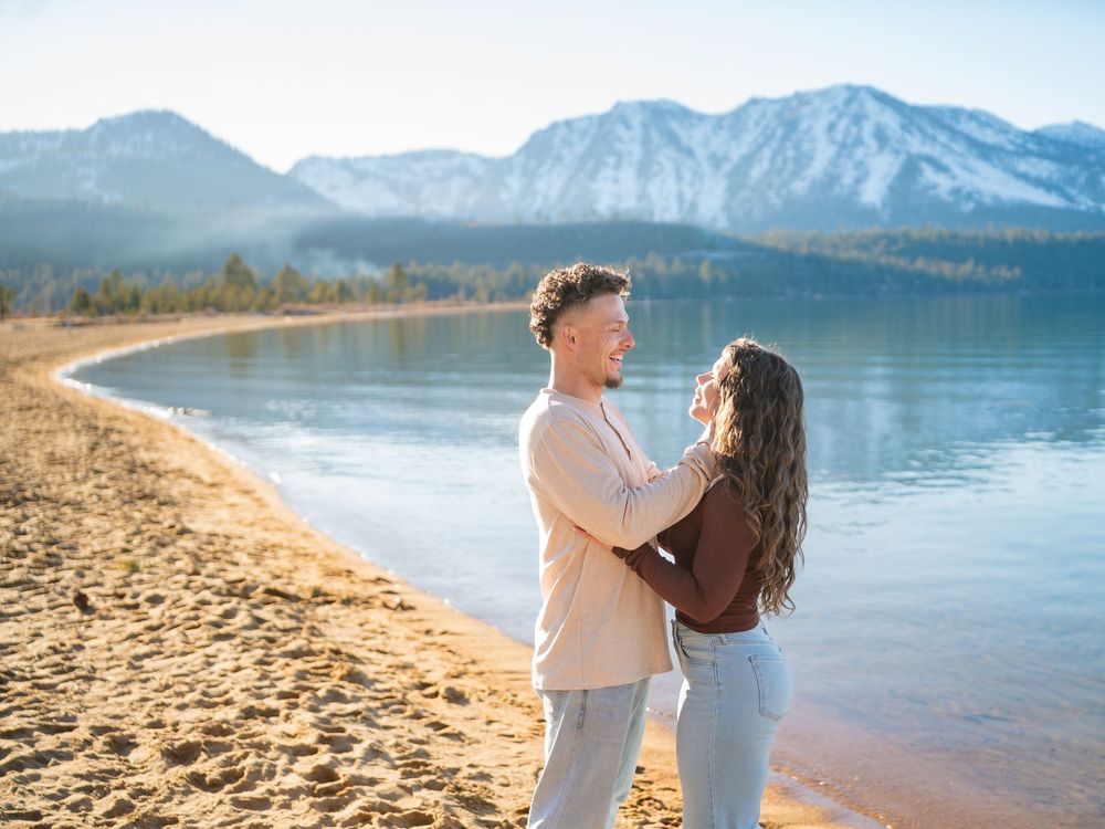 A couple shares a romantic moment on a sandy beach with mountains and lake in the background.
