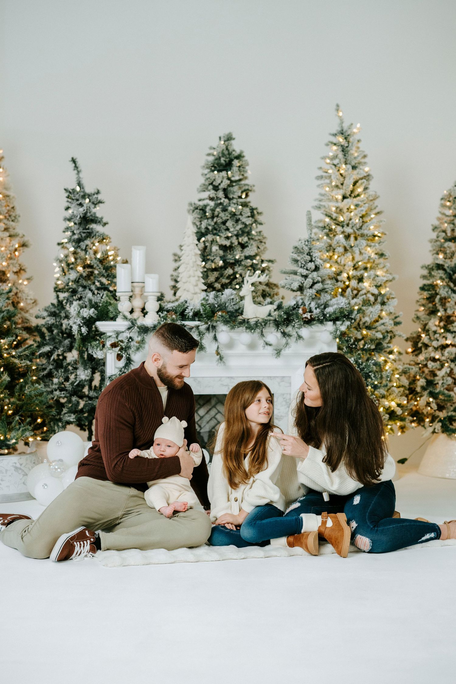 Family lounging together on white floor with decorated Christmas trees and mantel display behind them.