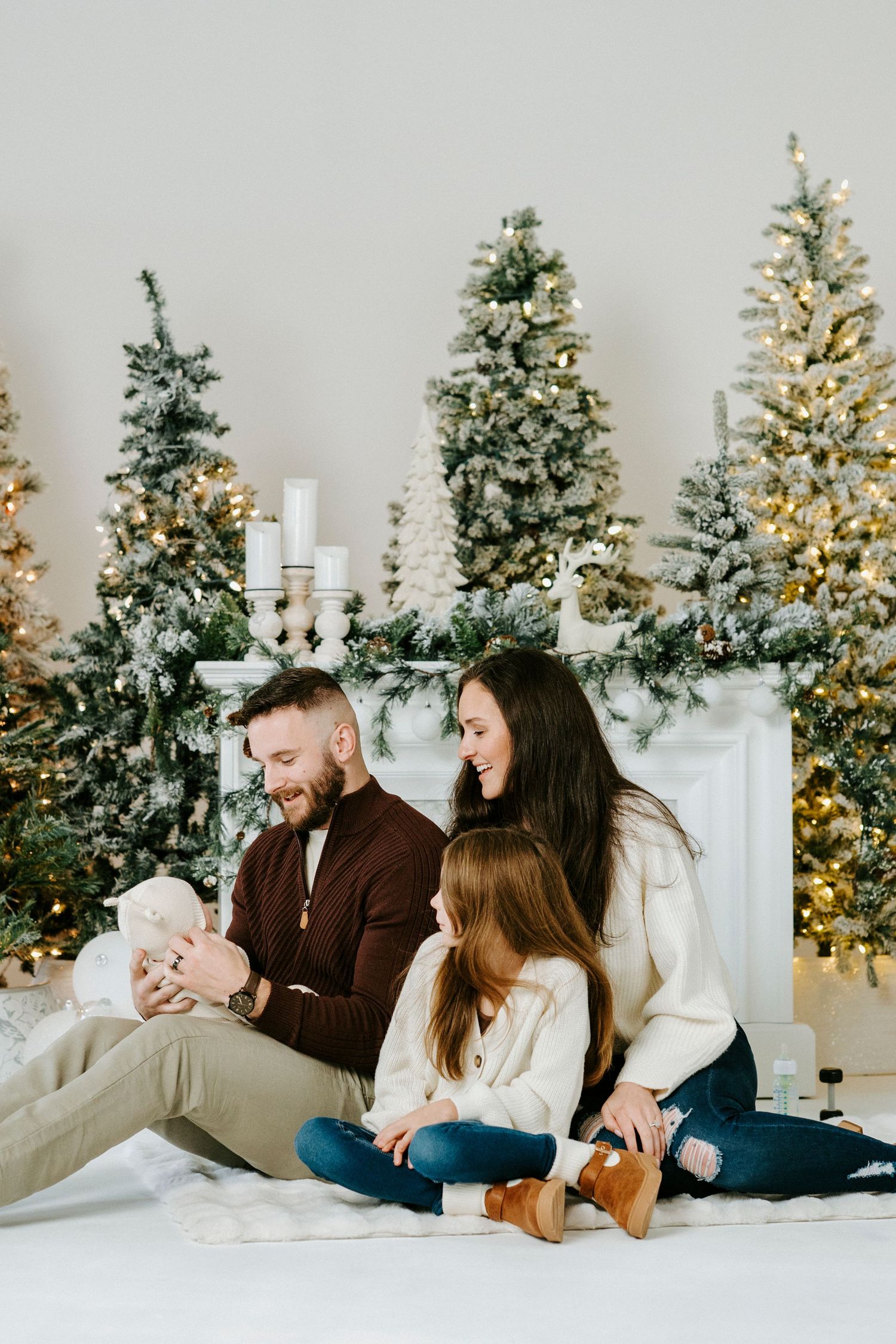 A family sits together in front of multiple lit Christmas trees and a decorated fireplace mantel during the holiday season.
