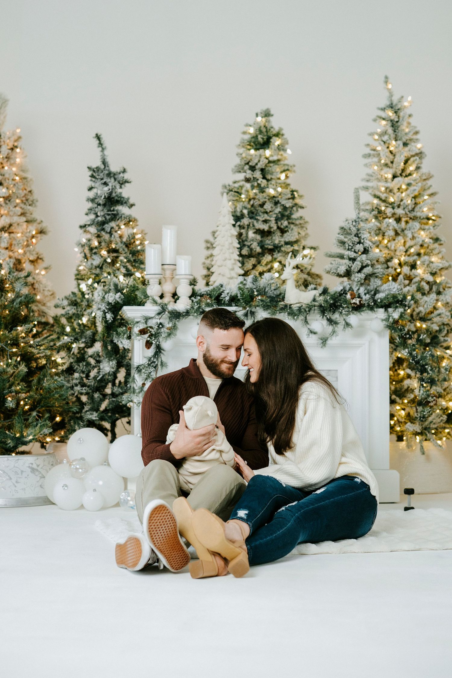 Cozy holiday scene with multiple decorated Christmas trees and white fireplace backdrop.