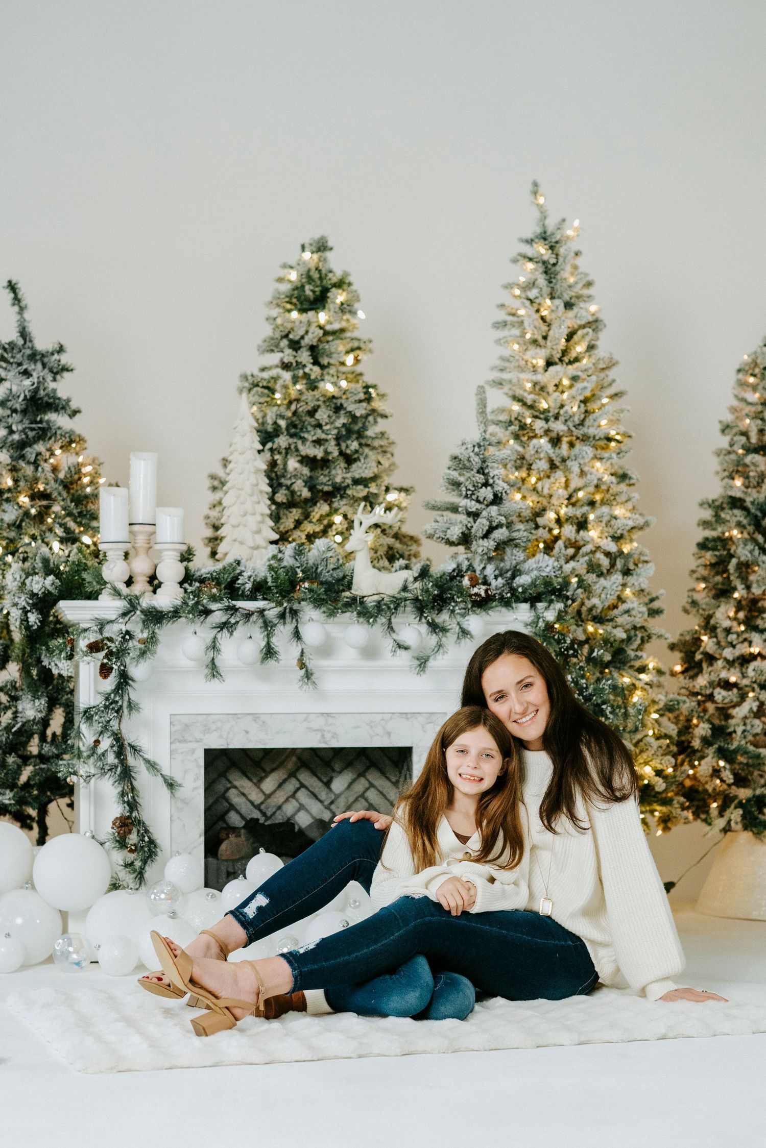 A cozy holiday scene with two people sitting on white blankets in front of decorated Christmas trees and a fireplace.