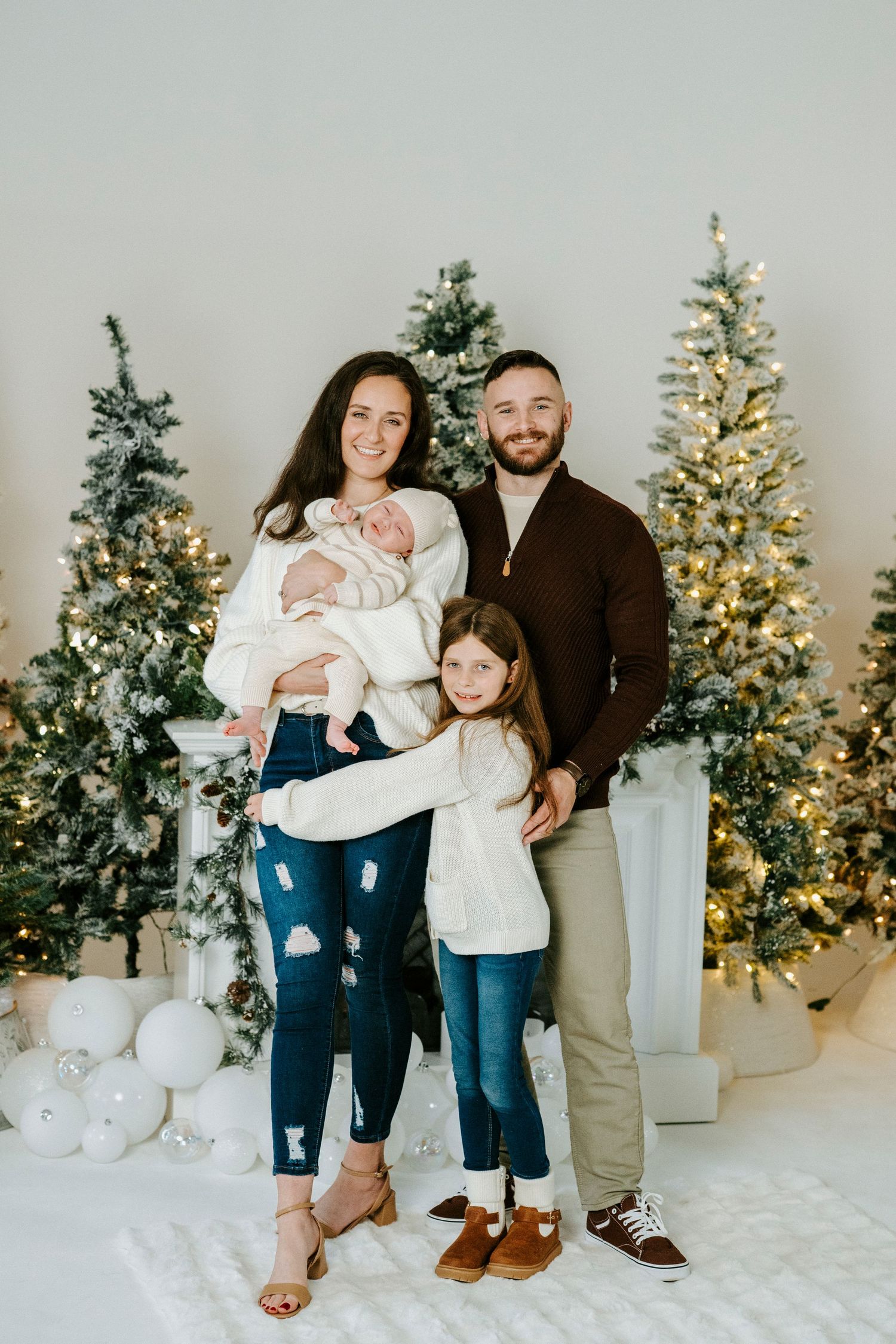 A family poses together in front of multiple lit Christmas trees in a bright white studio setting.
