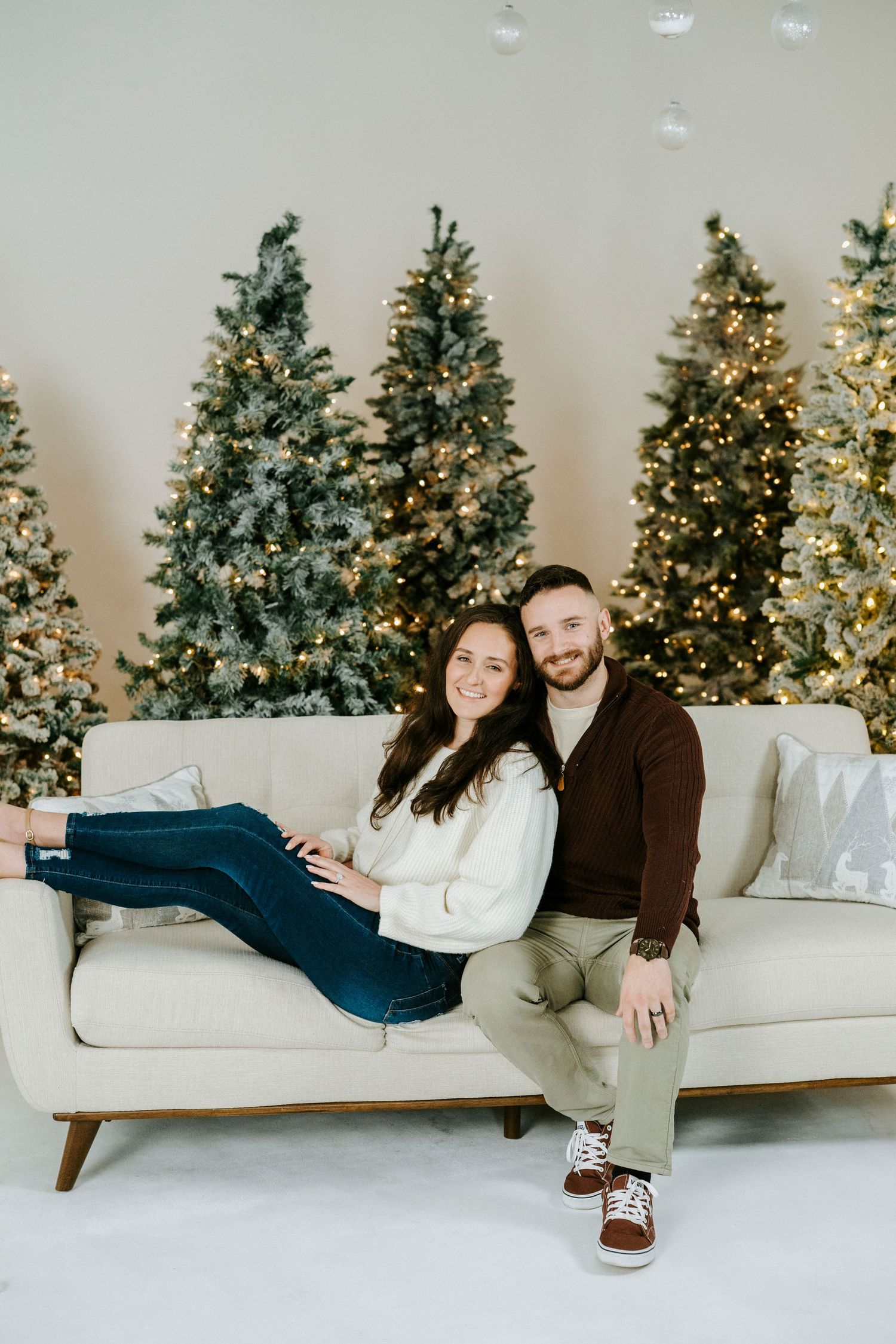 A couple relaxes on a white couch with Christmas trees glowing in the background.