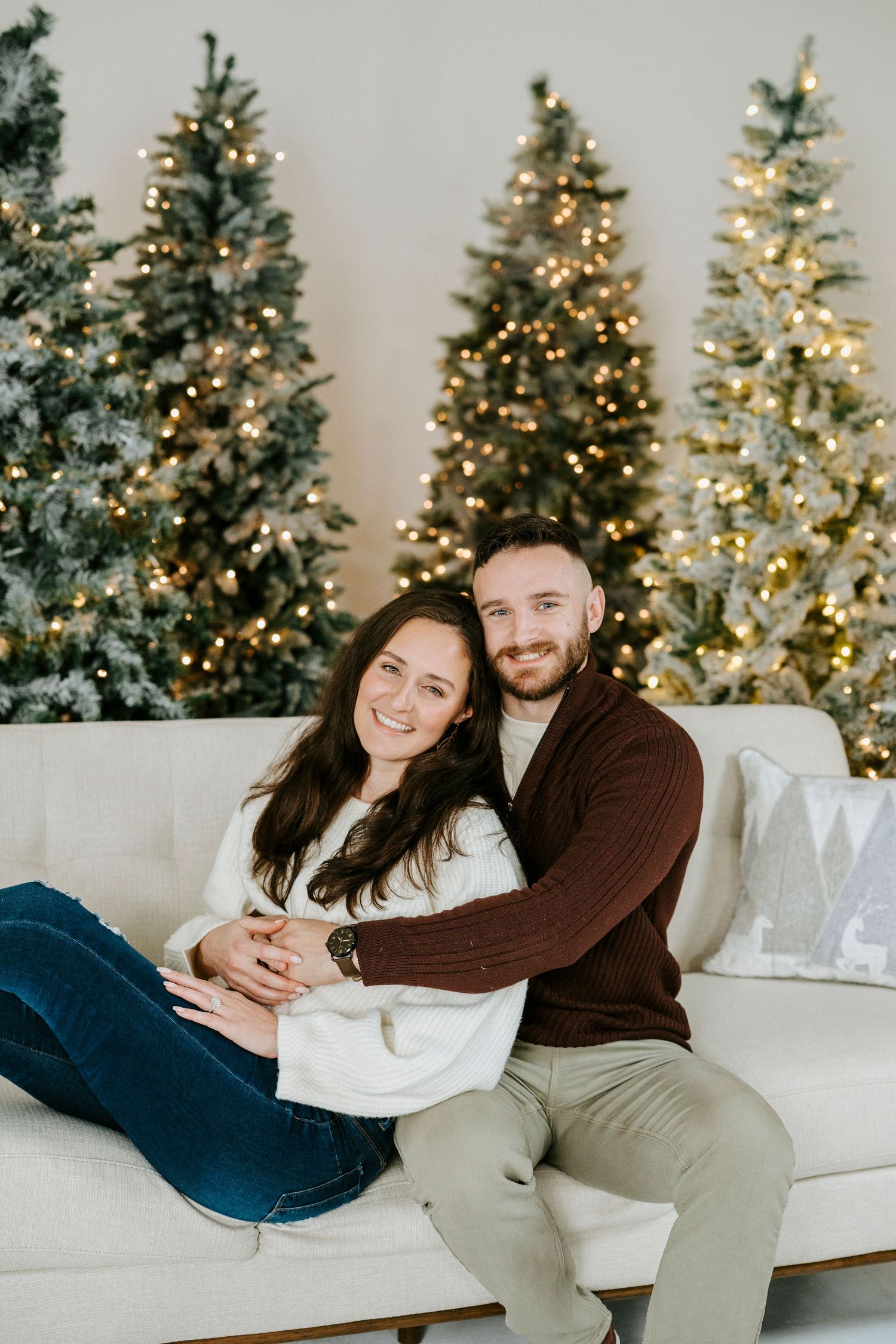 A couple relaxes on a white couch with illuminated Christmas trees creating a festive backdrop.