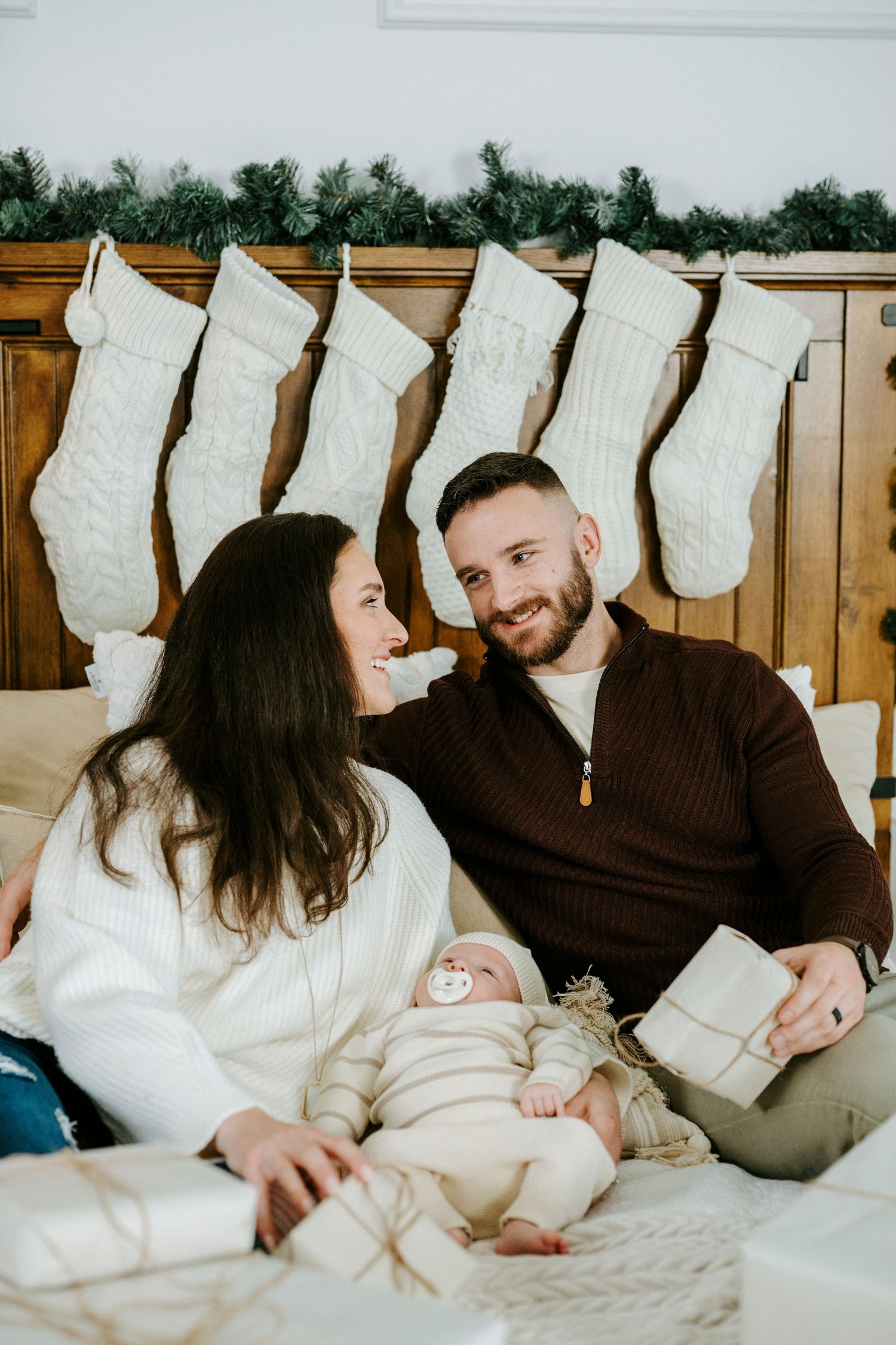 A family lounges together on a cozy bed with white stockings hanging on the wall behind them.