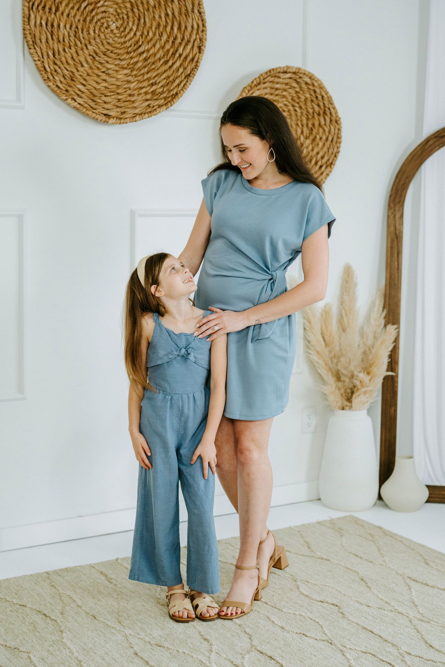 A parent and child wearing matching blue outfits share tender moments against a white wall with woven wall decor.