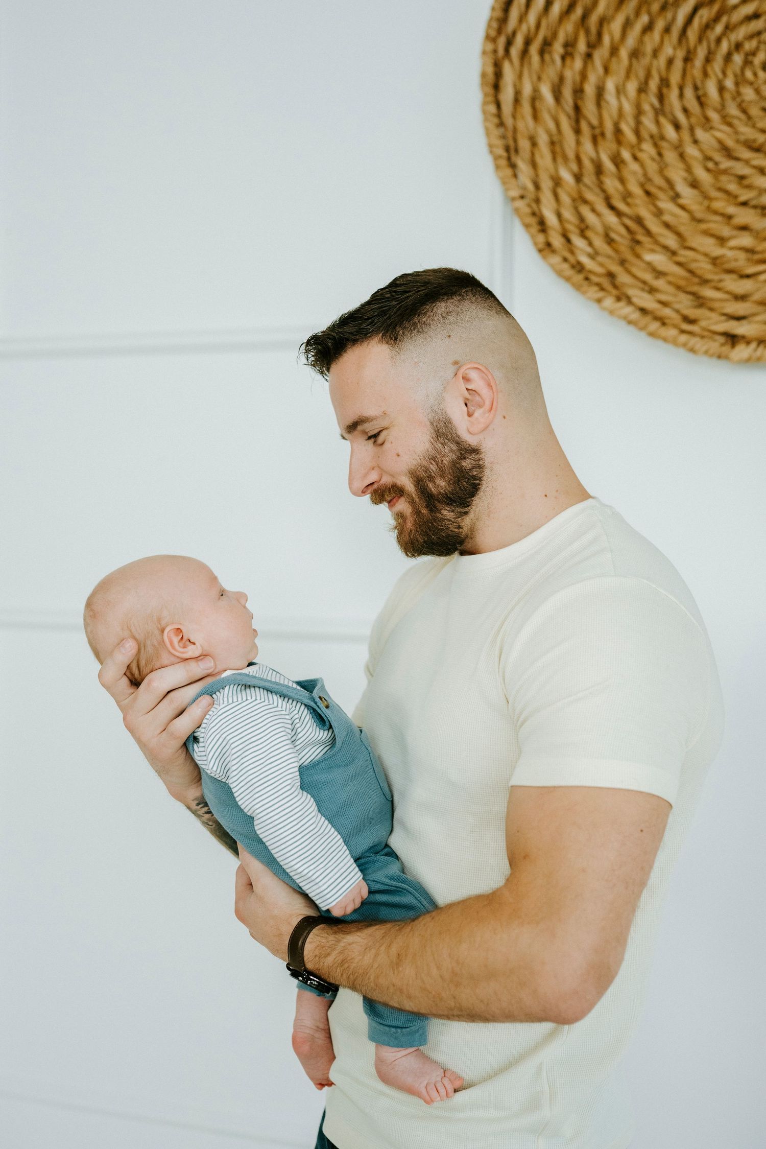 A tender family moment captured against a minimalist white backdrop with woven wall decorations.