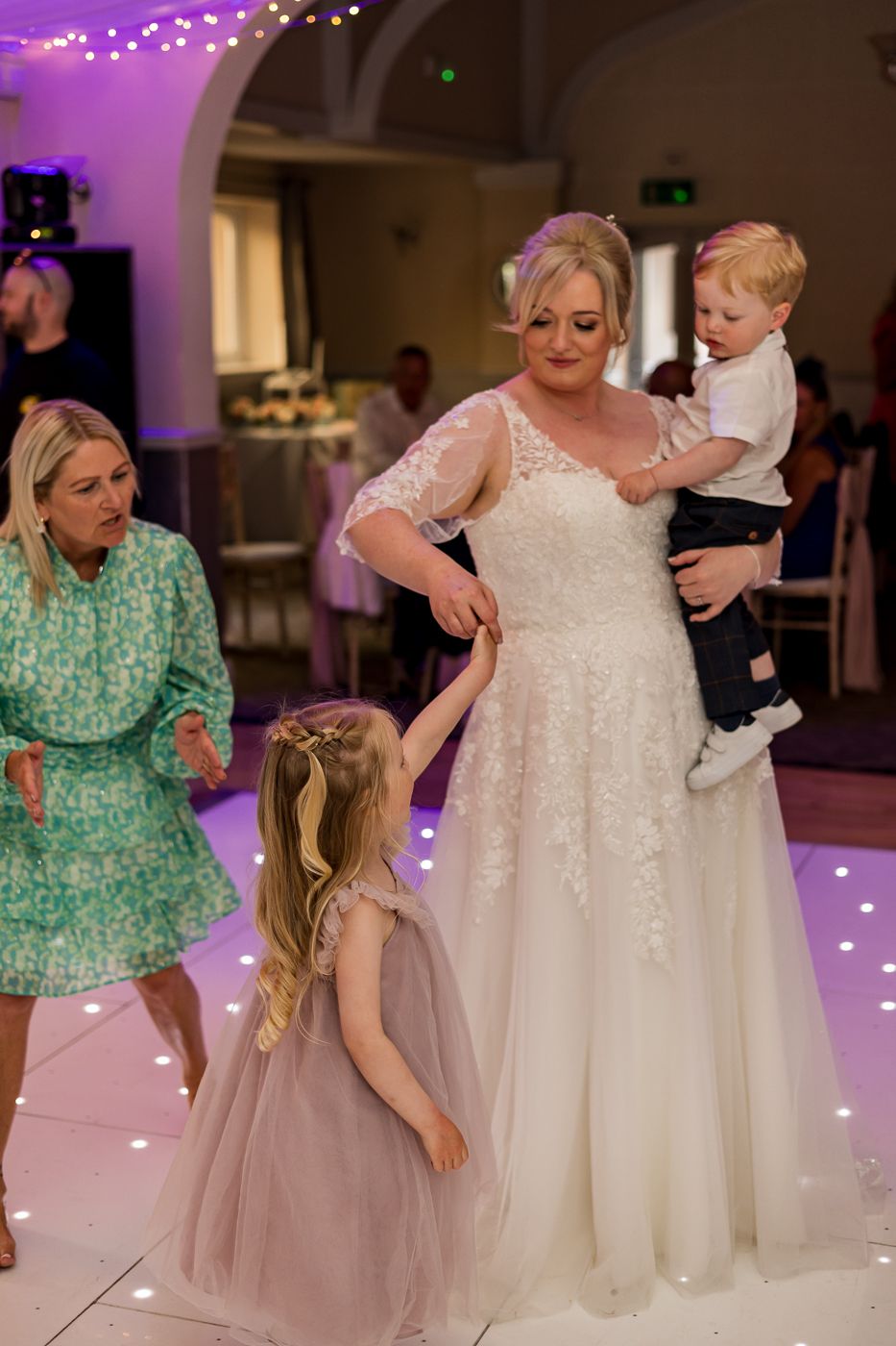 A bride in a lace gown dances with children on an illuminated dance floor at a wedding reception.
