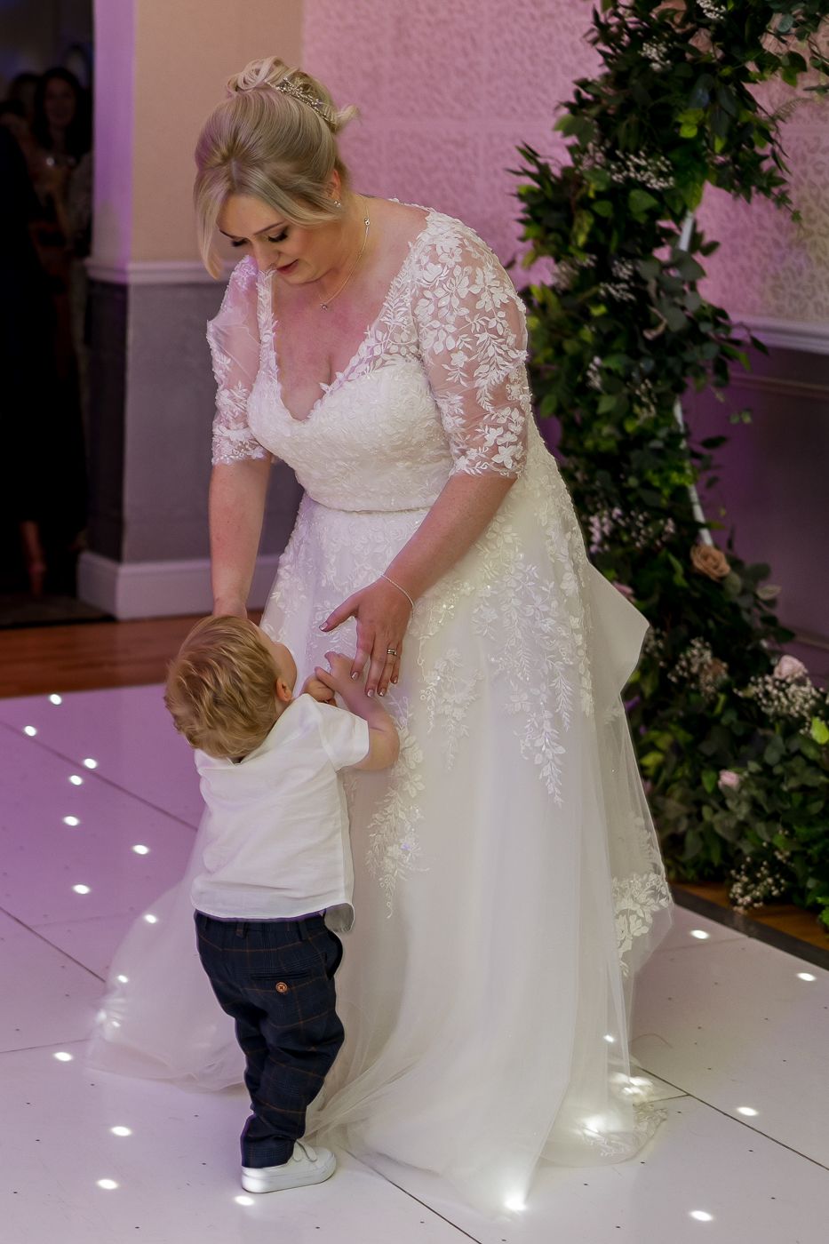 A bride in a lace wedding dress dances with a small child on an illuminated floor.