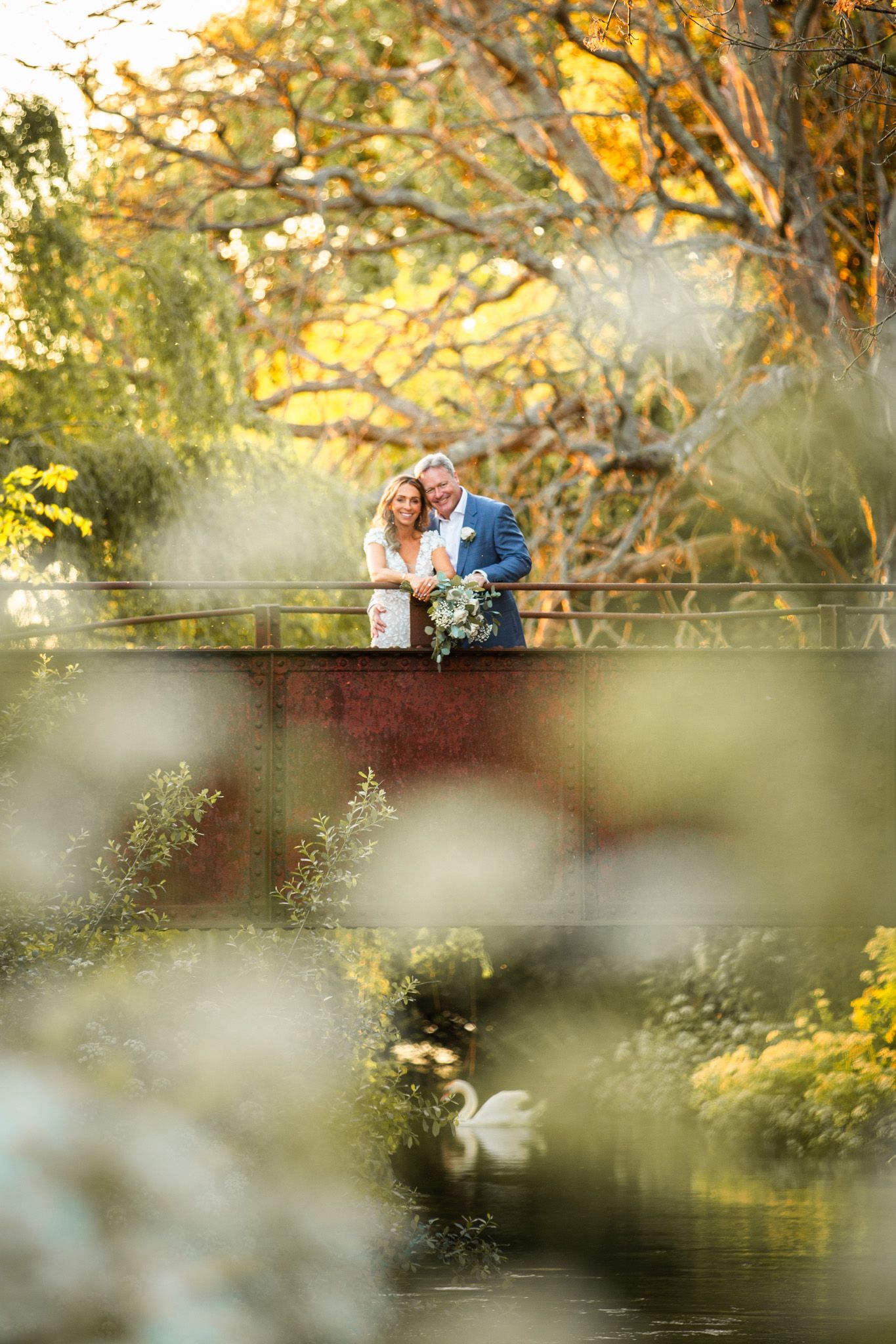A romantic couple stands on a wooden bridge overlooking a pond with autumn trees and golden sunlight in the background.