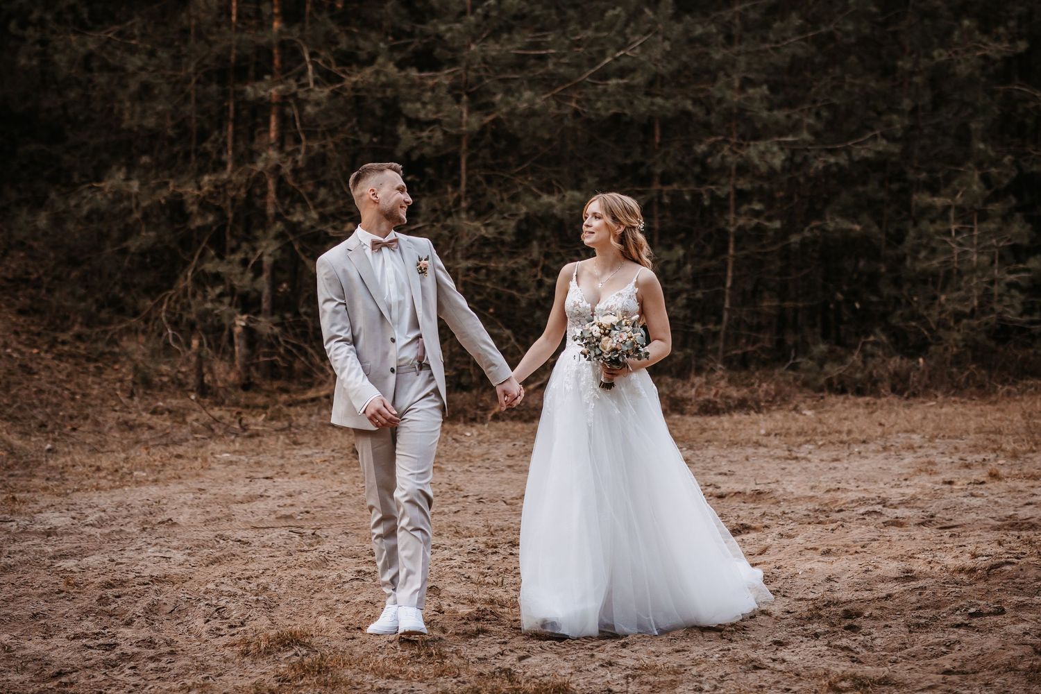 Newlyweds walking hand in hand through a forest clearing in elegant wedding attire.