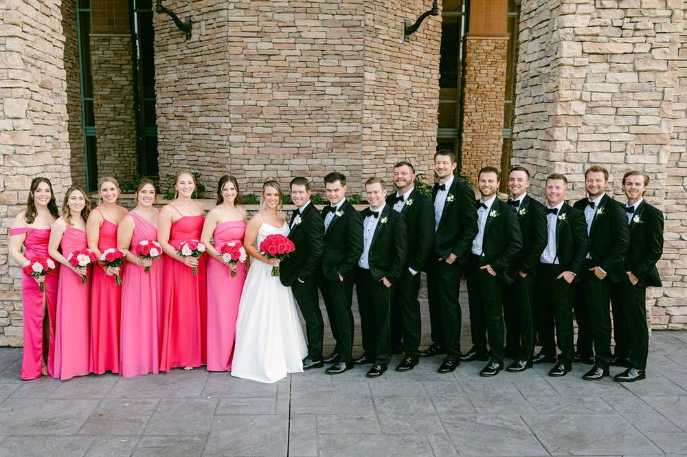 Wedding party in formal attire lined up against stone wall with bridesmaids in bright pink dresses and groomsmen in black suits.