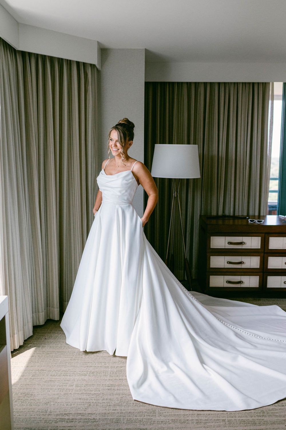 A flowing white wedding dress with a long train is displayed in a hotel room with green curtains.