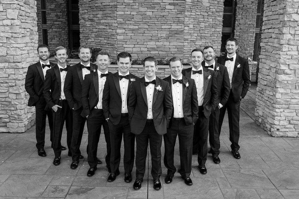 Groomsmen in black tuxedos pose in a V-formation against a stone wall backdrop.