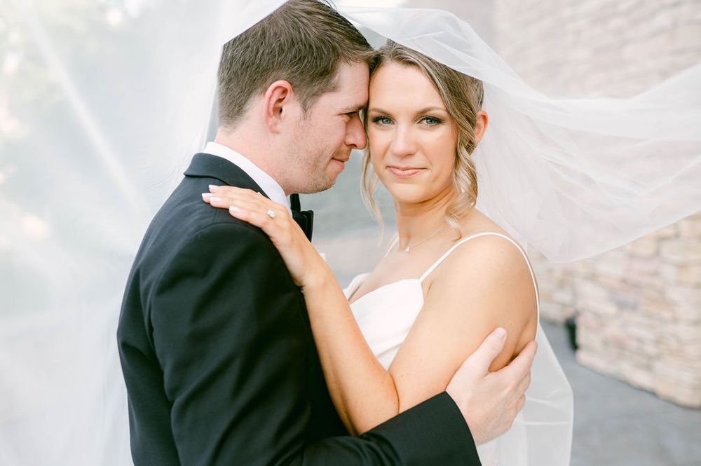 A romantic wedding portrait of a couple embracing against a light stone wall with soft lighting.
