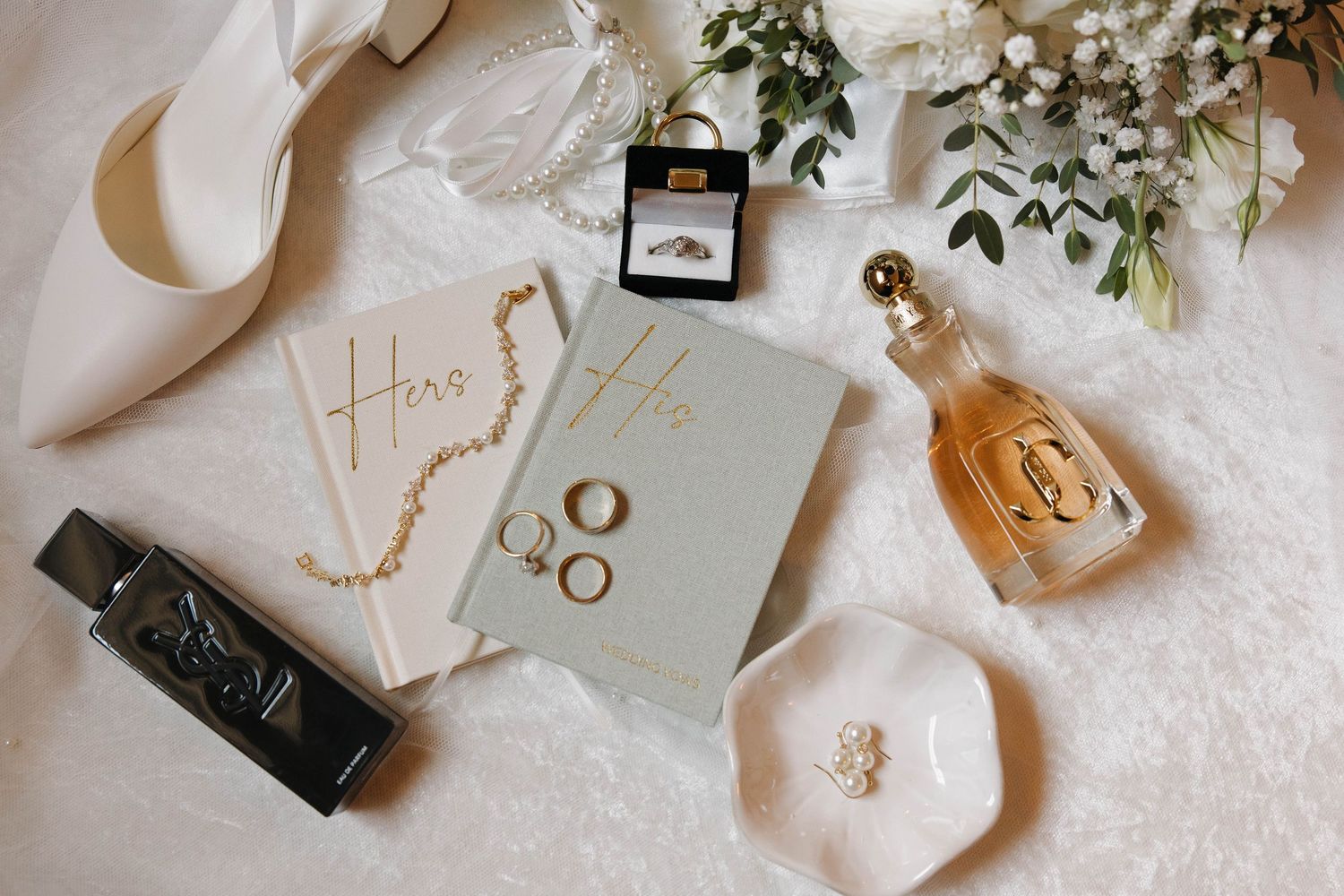 Wedding details including rings, invitations, shoes, and floral decor arranged on white textured surface in soft lighting.