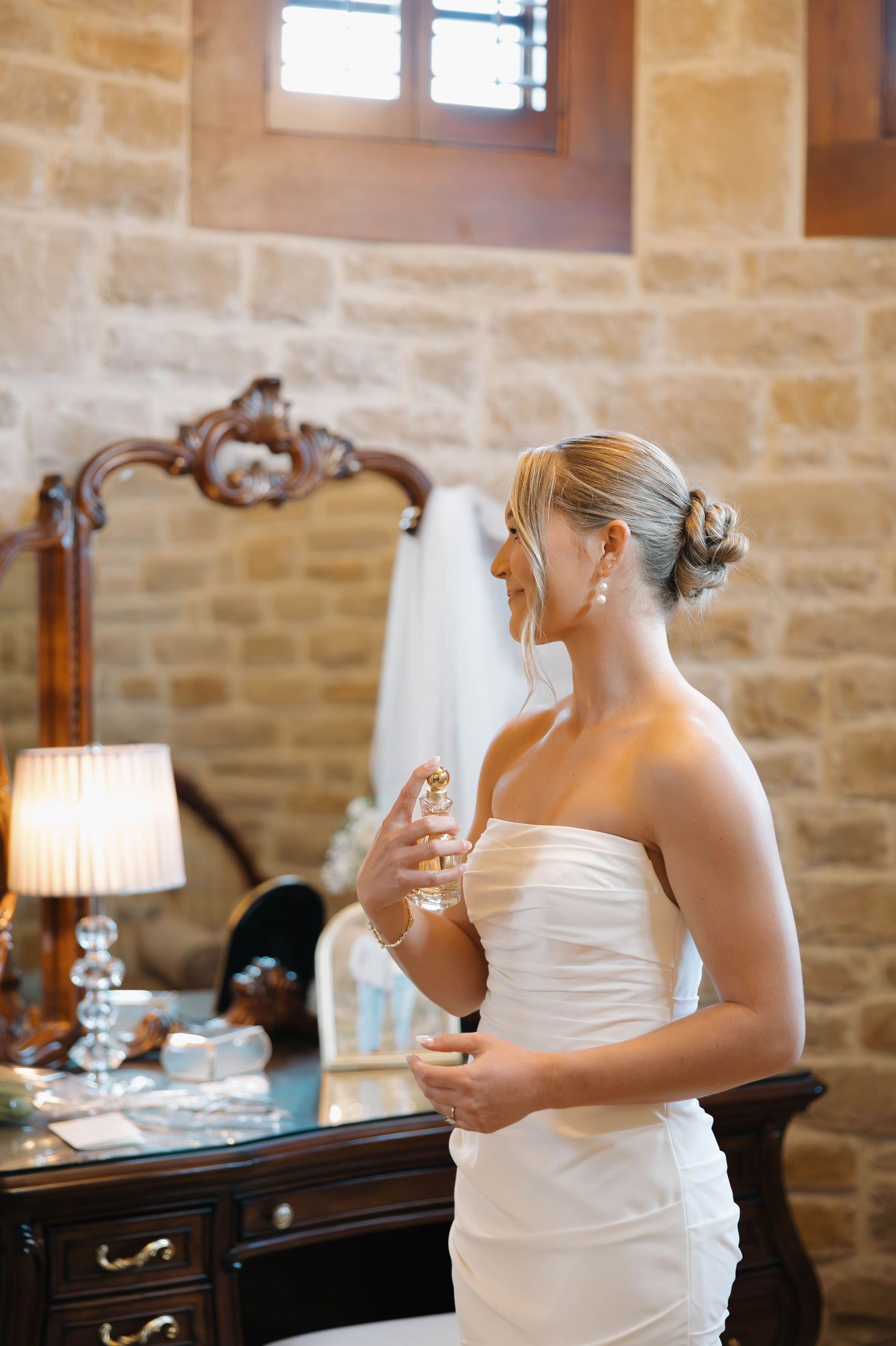 Bride in a strapless white gown applies perfume while standing in a stone-walled room.