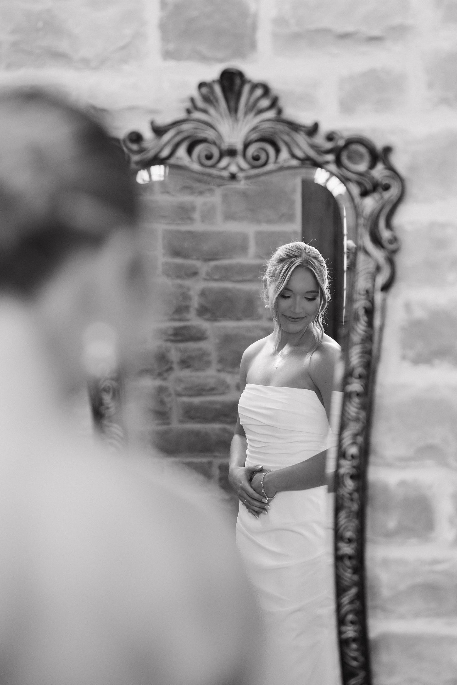 A bride in a white dress looks at her reflection in an ornate mirror during wedding preparations.