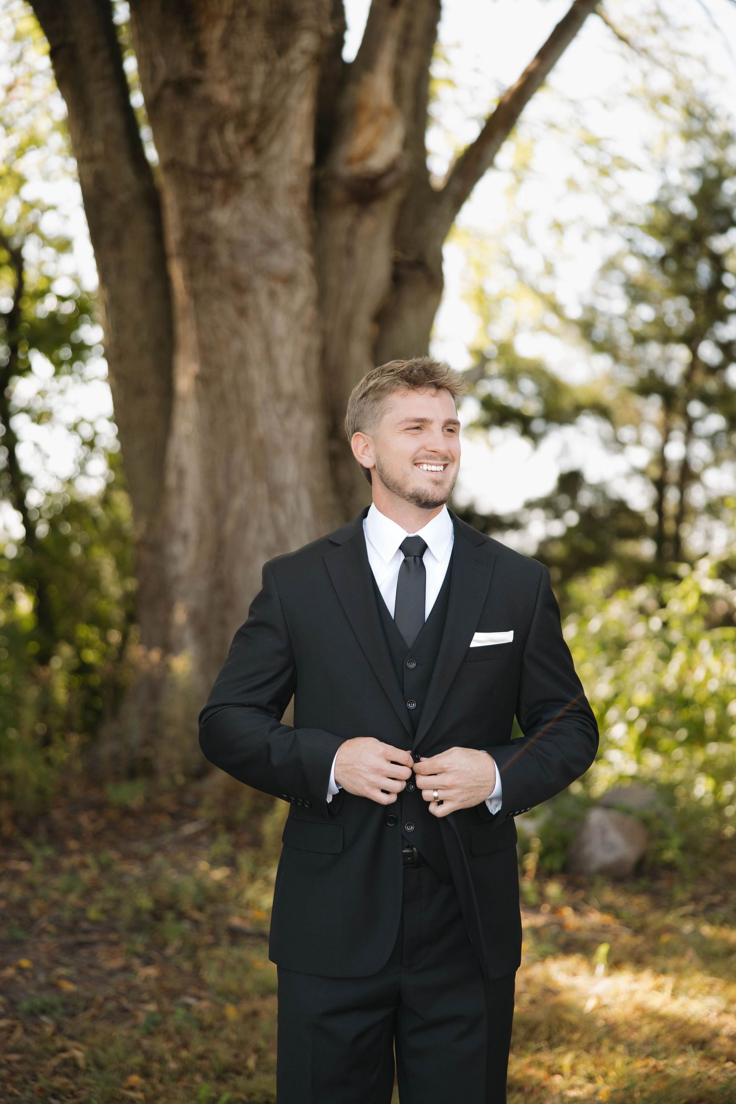 A groom in a classic black tuxedo stands in a wooded outdoor setting during golden hour.