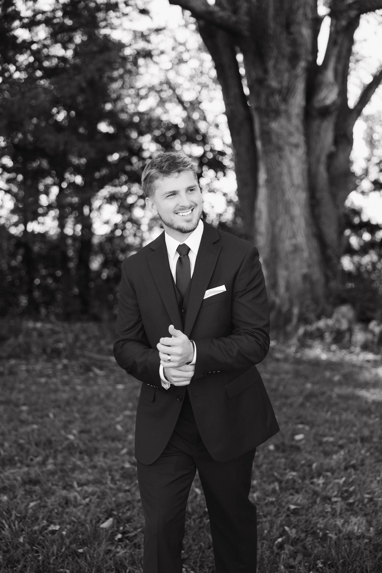 A groom in a classic black suit and tie poses outdoors against a backdrop of trees in a black and white portrait.