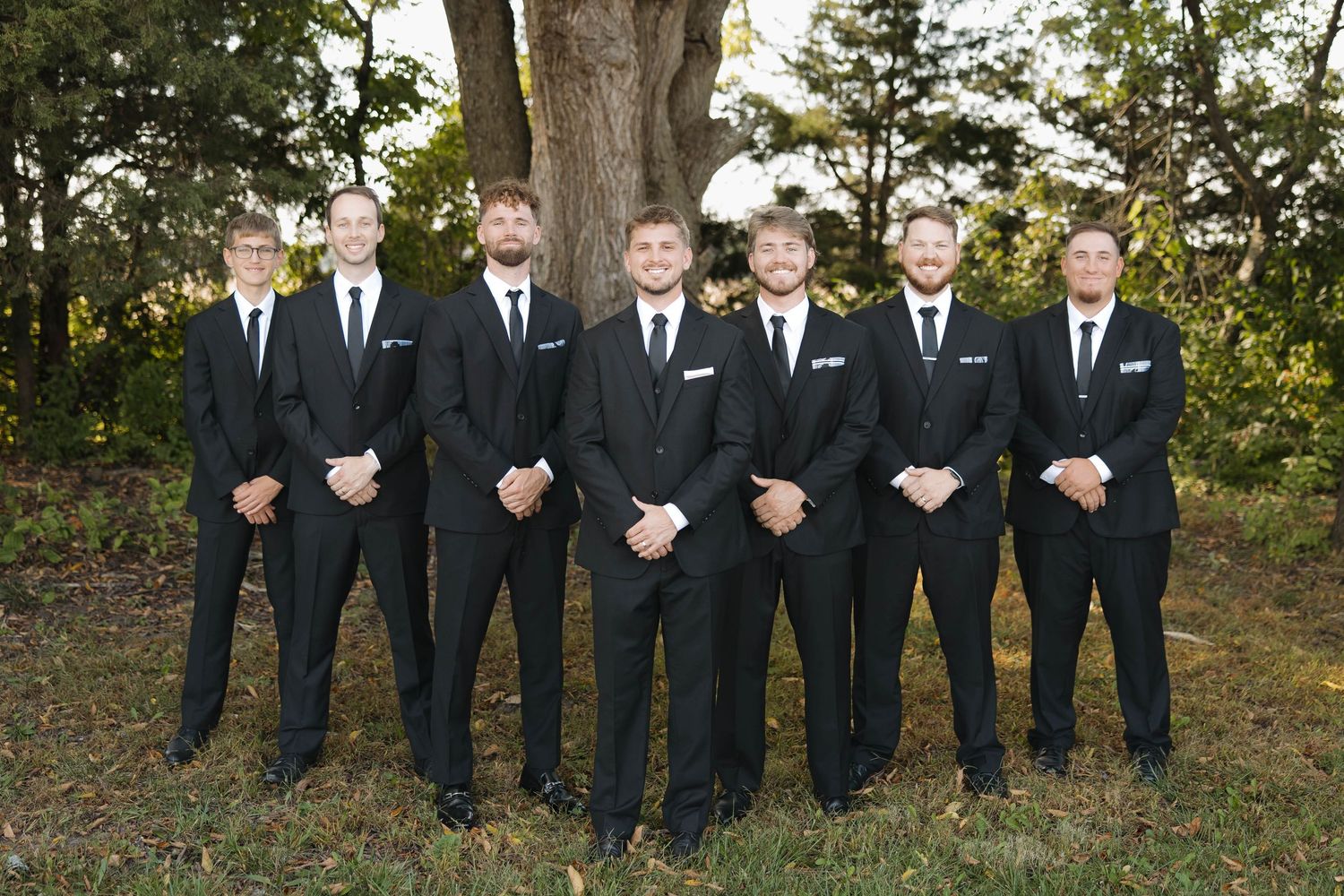 A group of groomsmen in black suits stand in a row outdoors against a wooded background.
