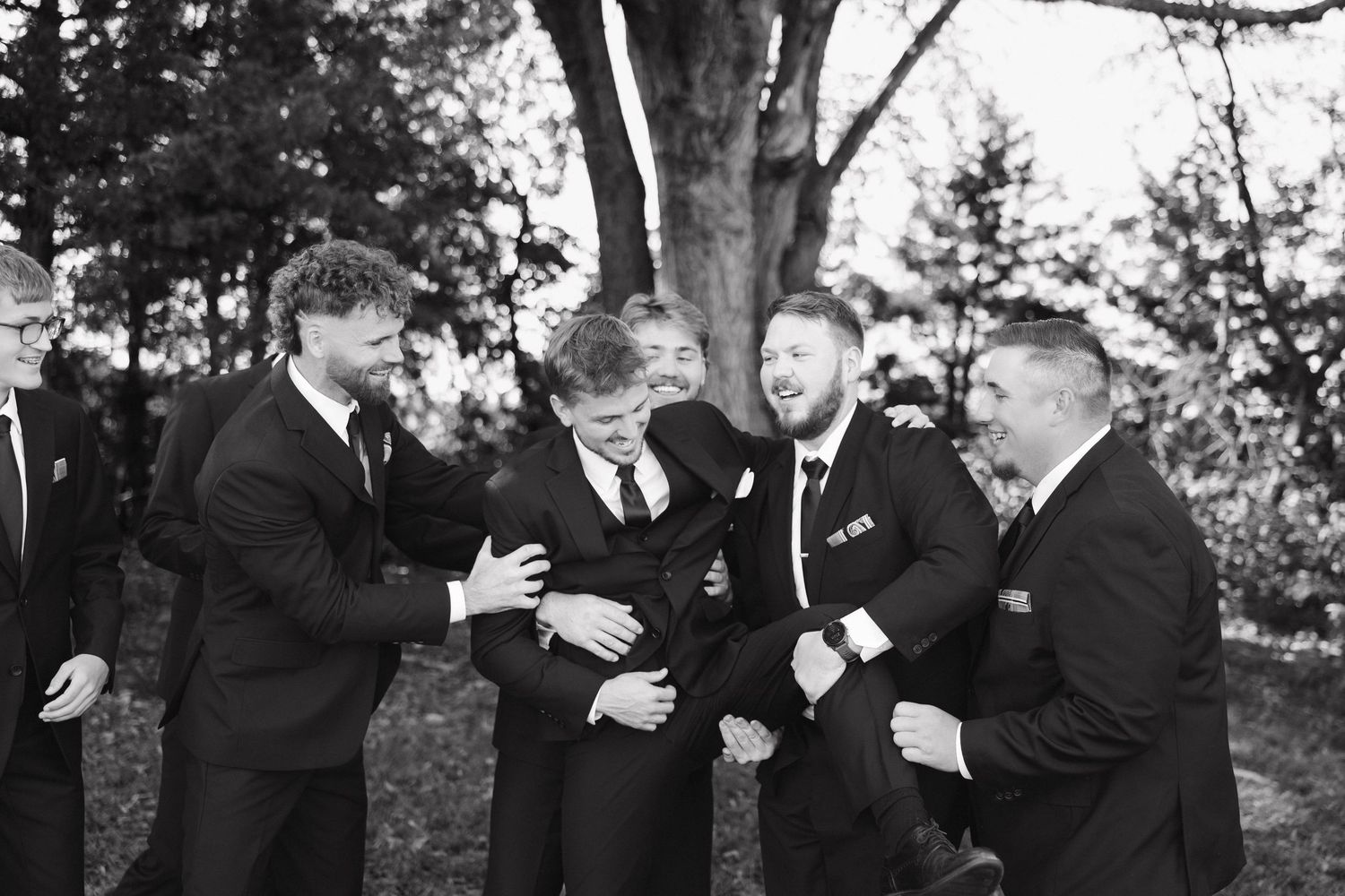 A sequence of black and white photos showing groomsmen in suits sharing emotional reactions and embraces outdoors.