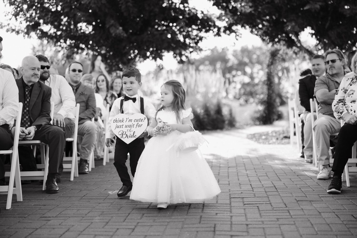 Wedding ceremony processional on a brick paved pathway with guests seated on white chairs.