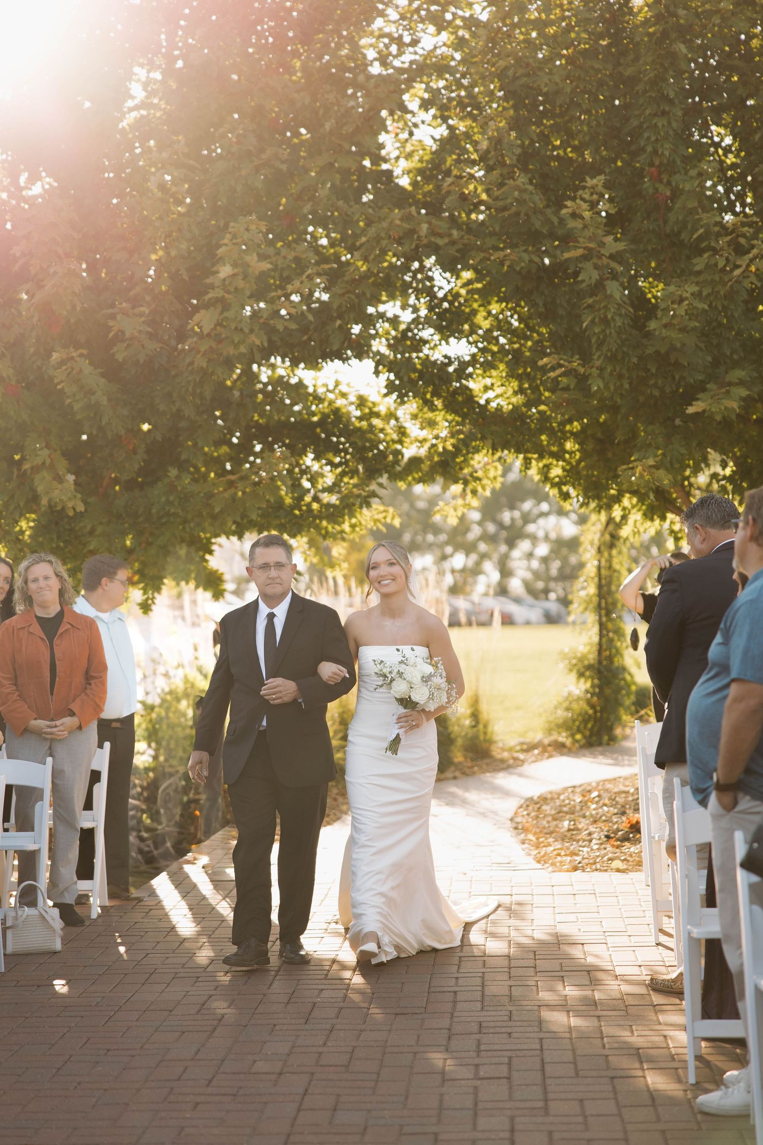 Bride in white gown walks down brick path during outdoor ceremony processional.