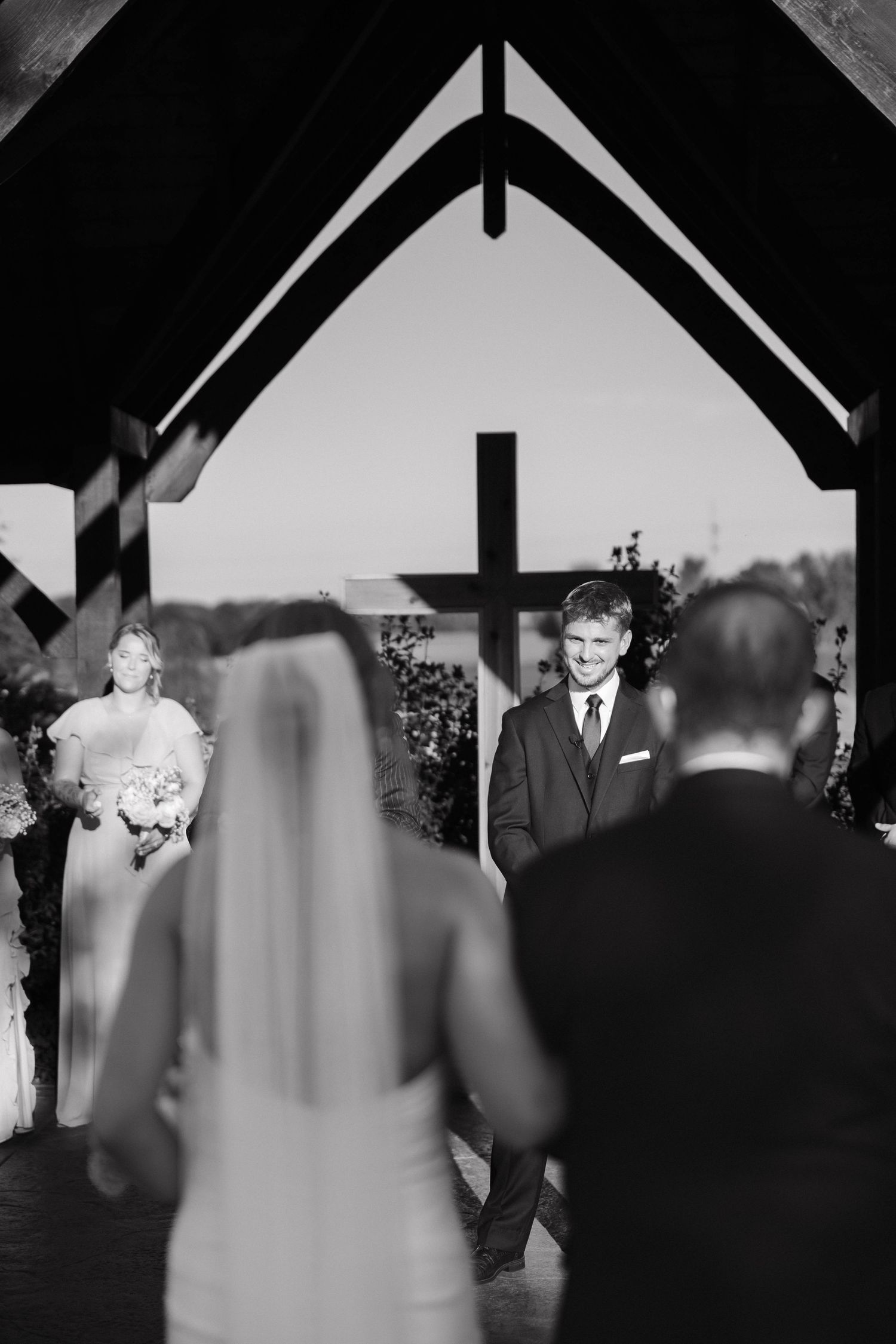 Black and white photo of a wedding ceremony under a wooden chapel arch with a cross.