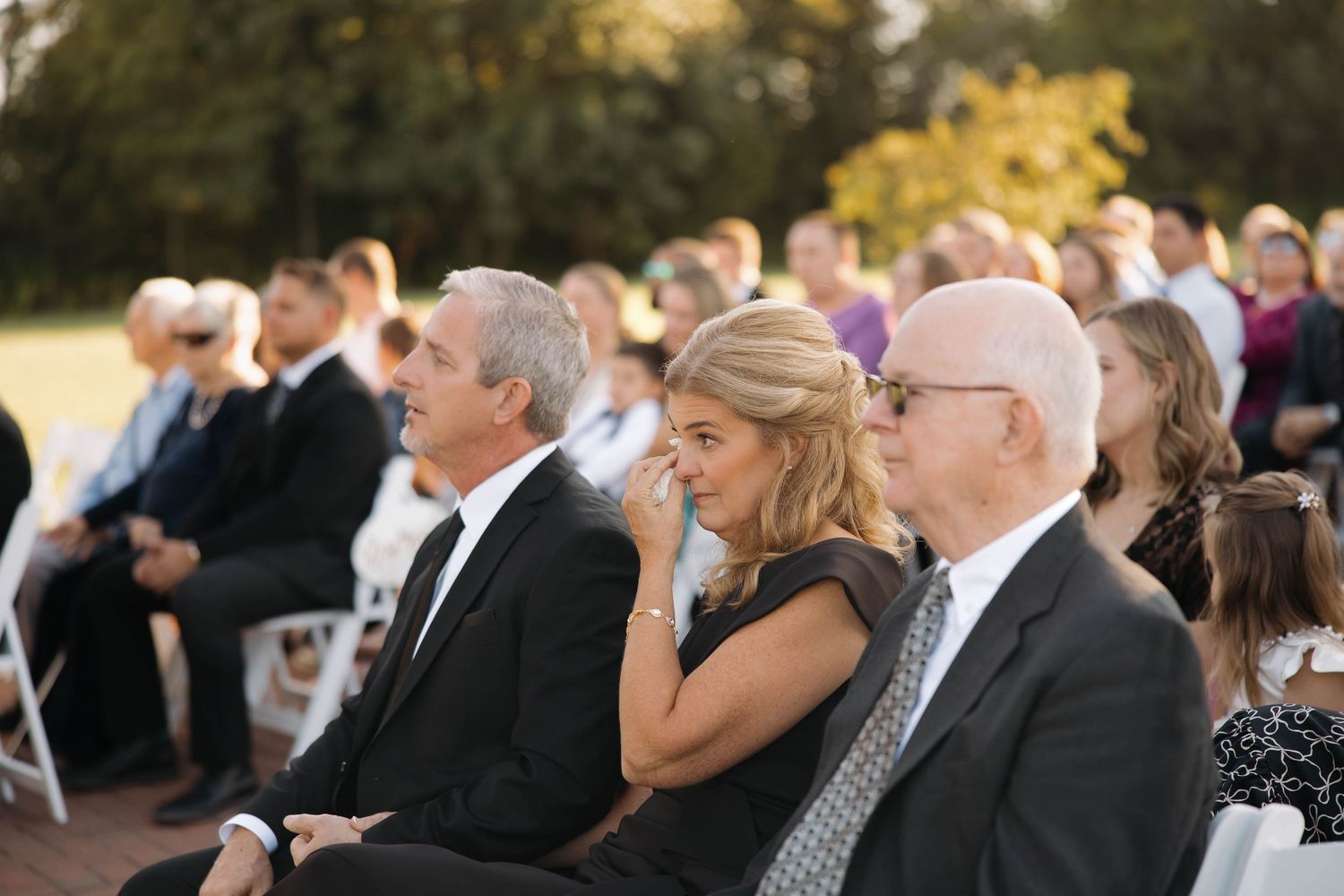 Wedding guests seated in rows outdoors during a ceremony with autumn foliage in the background.