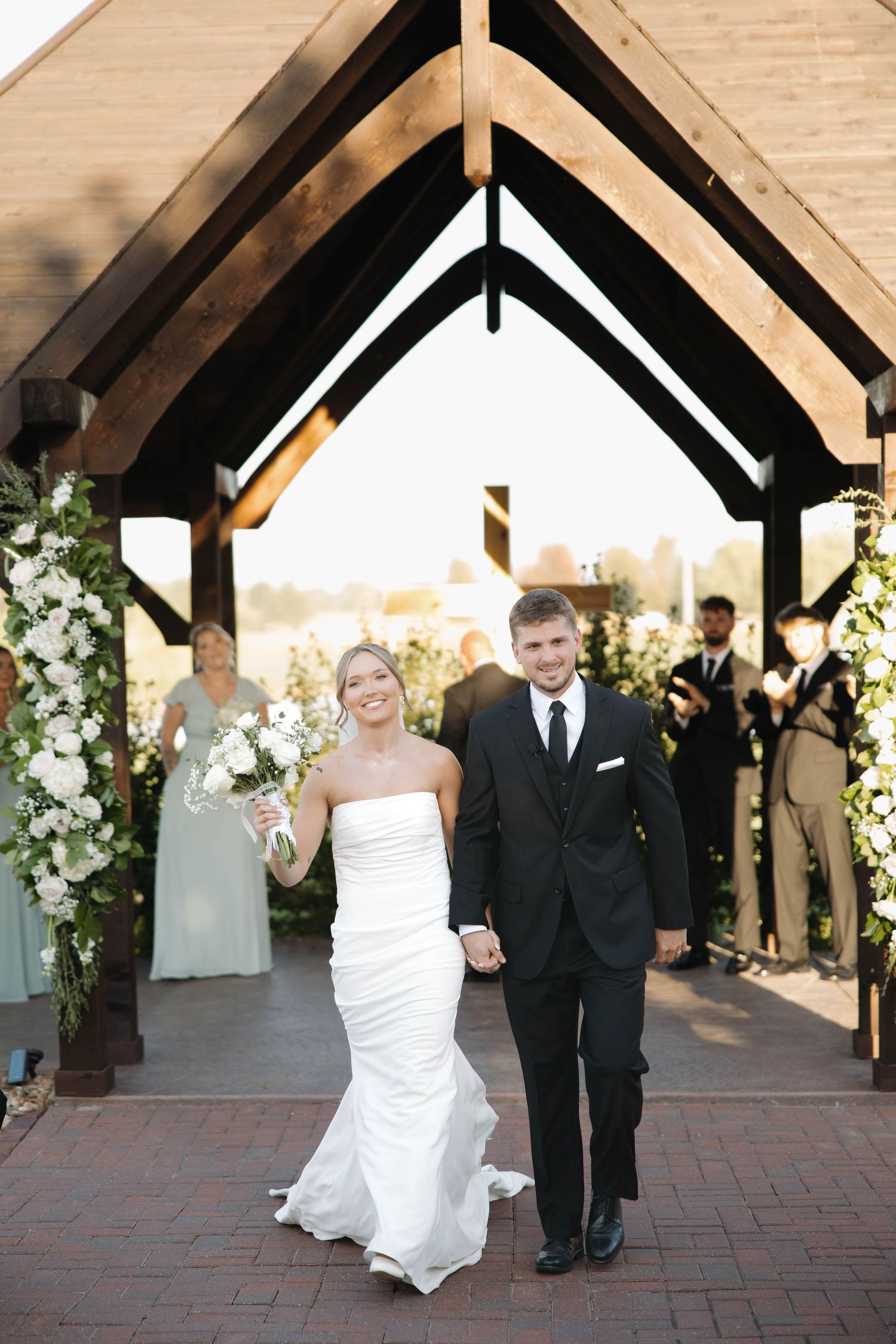 Newlyweds exit their wedding ceremony under a wooden archway decorated with white floral arrangements.