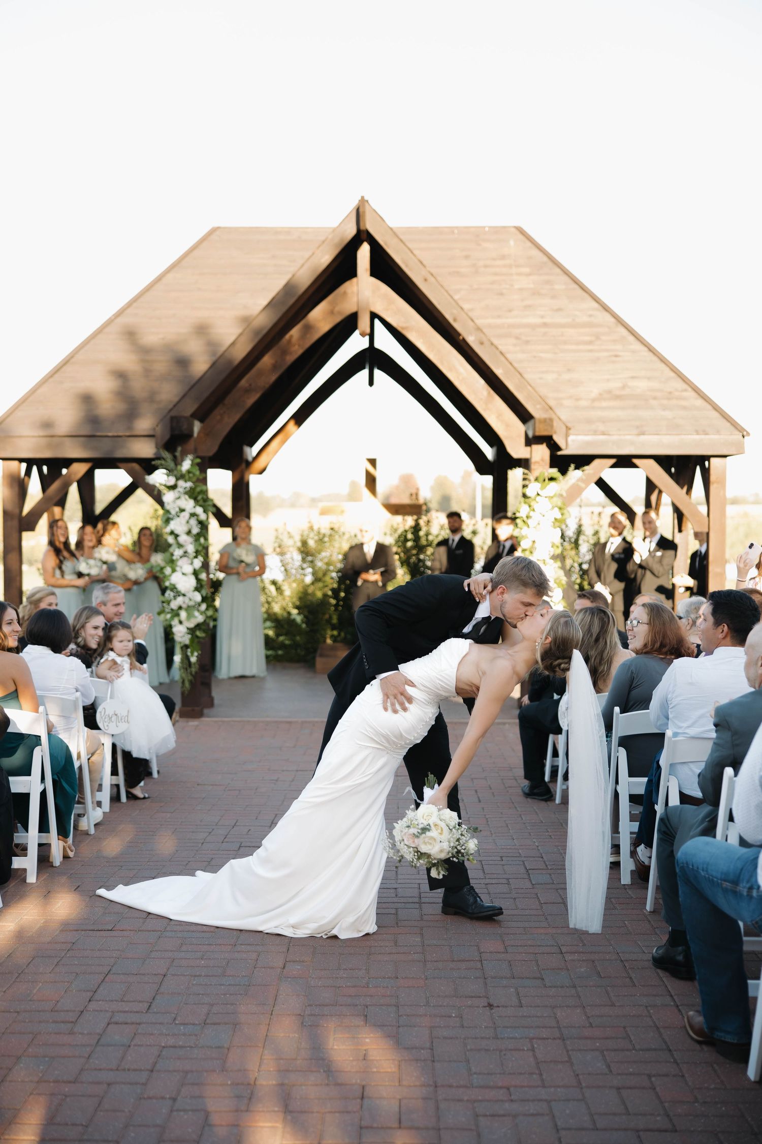 Wedding guests celebrate under wooden gazebos during an outdoor ceremony on brick pavement.