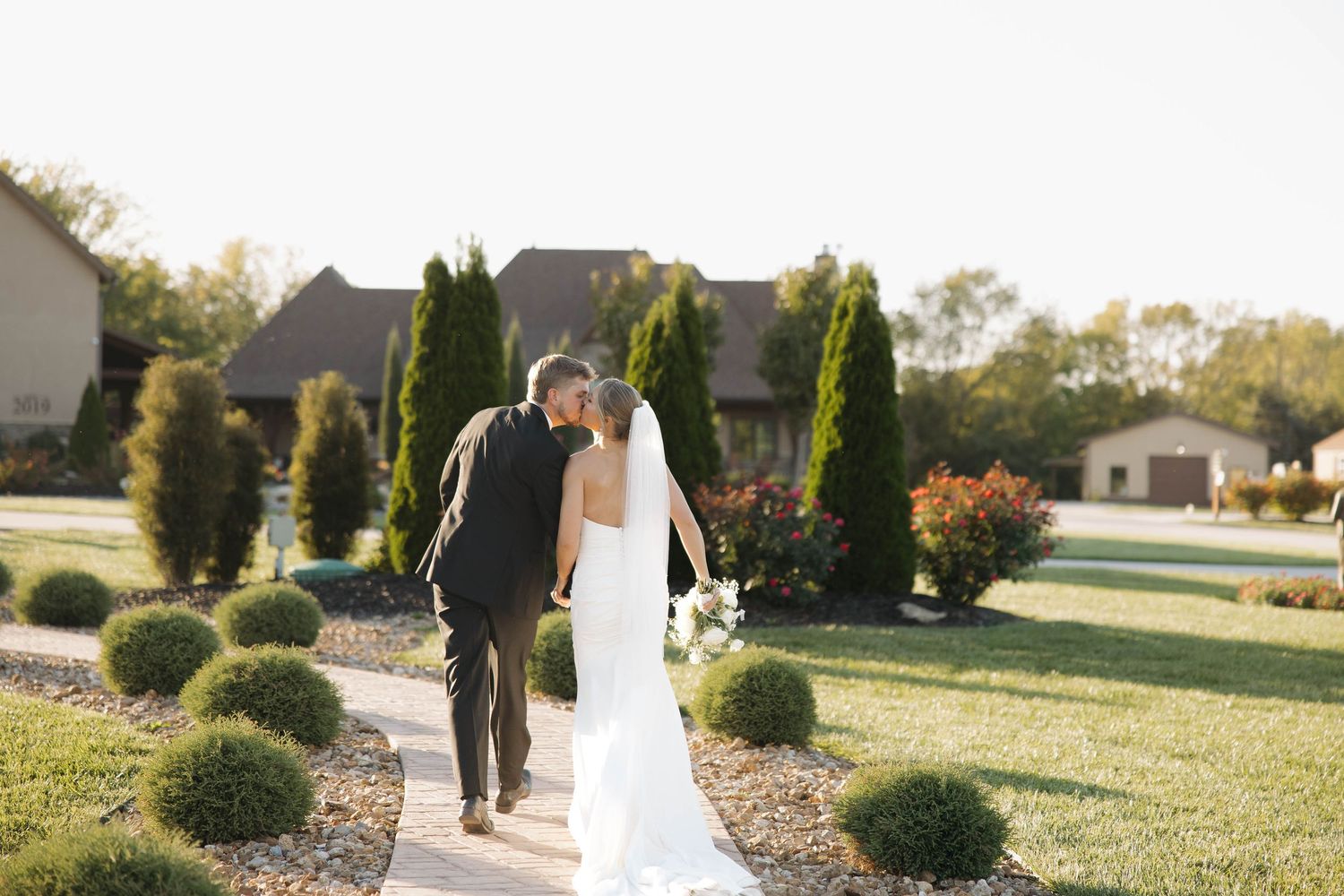 Wedding party walks along a landscaped garden path with evergreen shrubs and autumn foliage.
