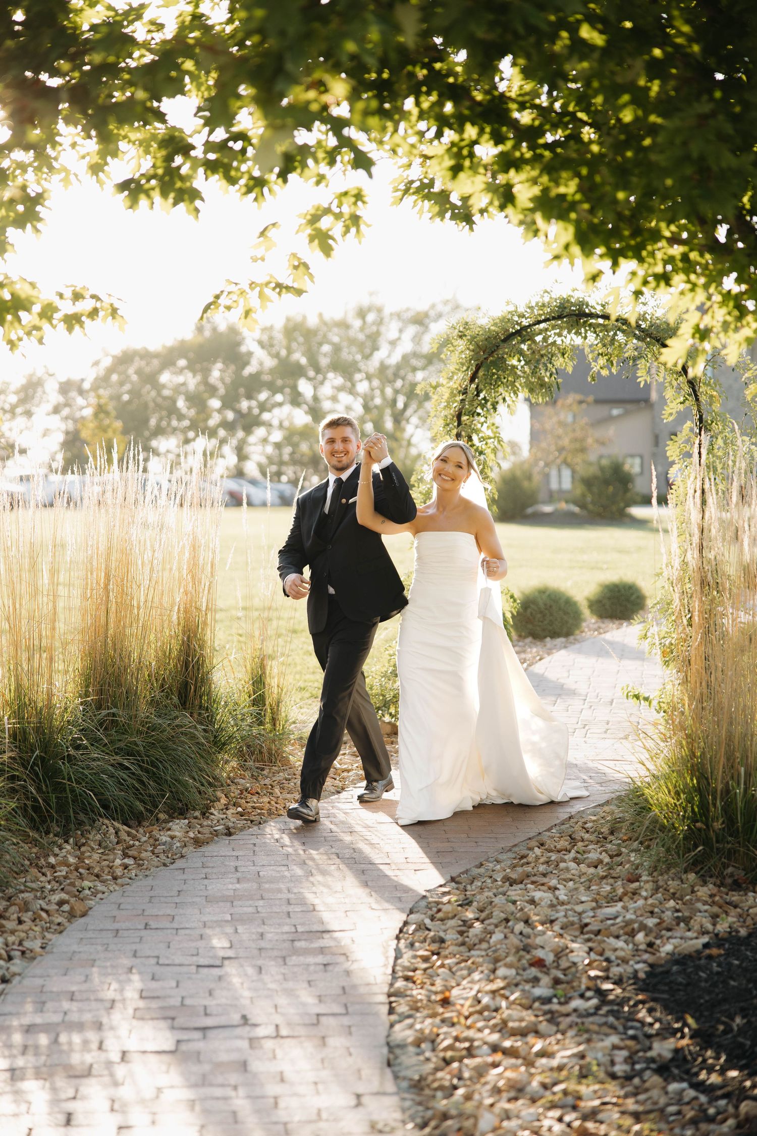 Couple walks along a winding stone pathway surrounded by ornamental grasses in golden sunlight.