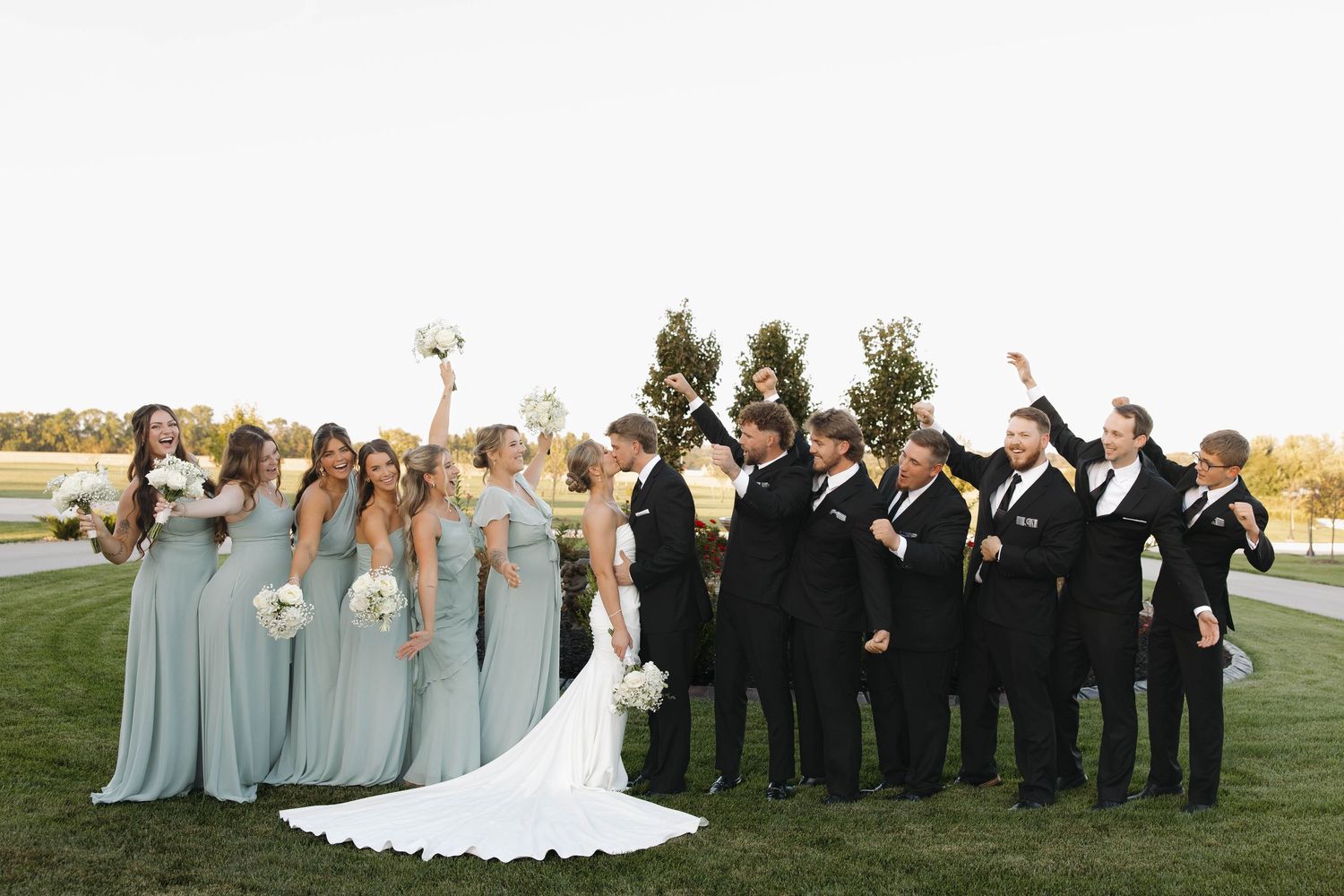Wedding party in black suits and light blue dresses poses together outdoors at sunset.