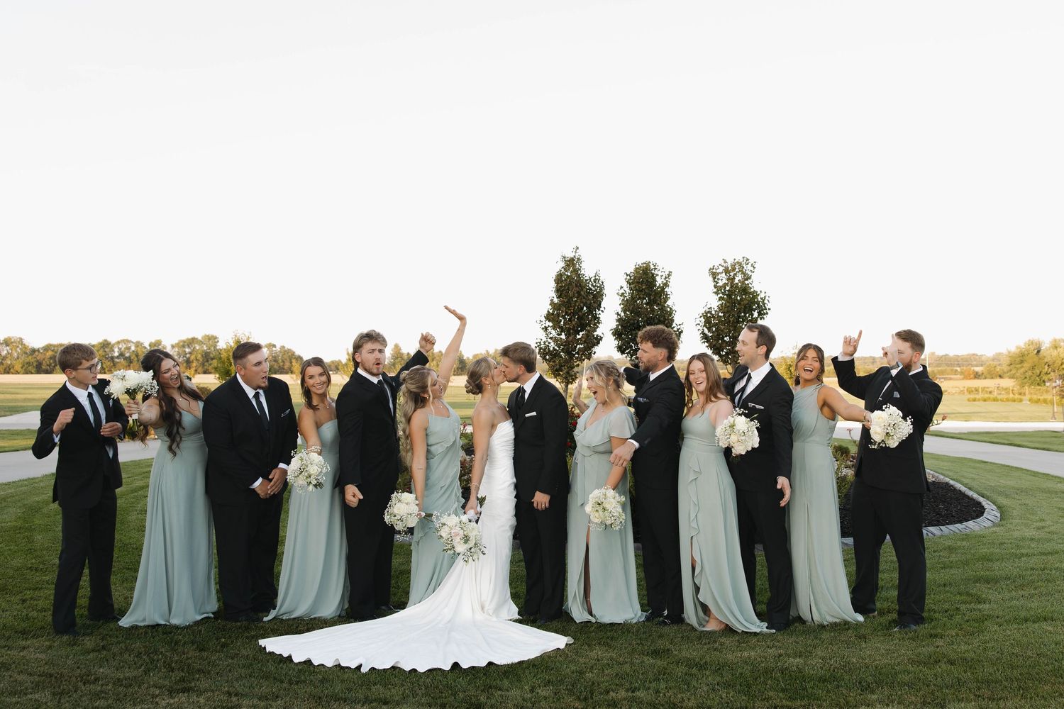 Wedding party in light blue dresses and black tuxedos poses together on a manicured lawn.