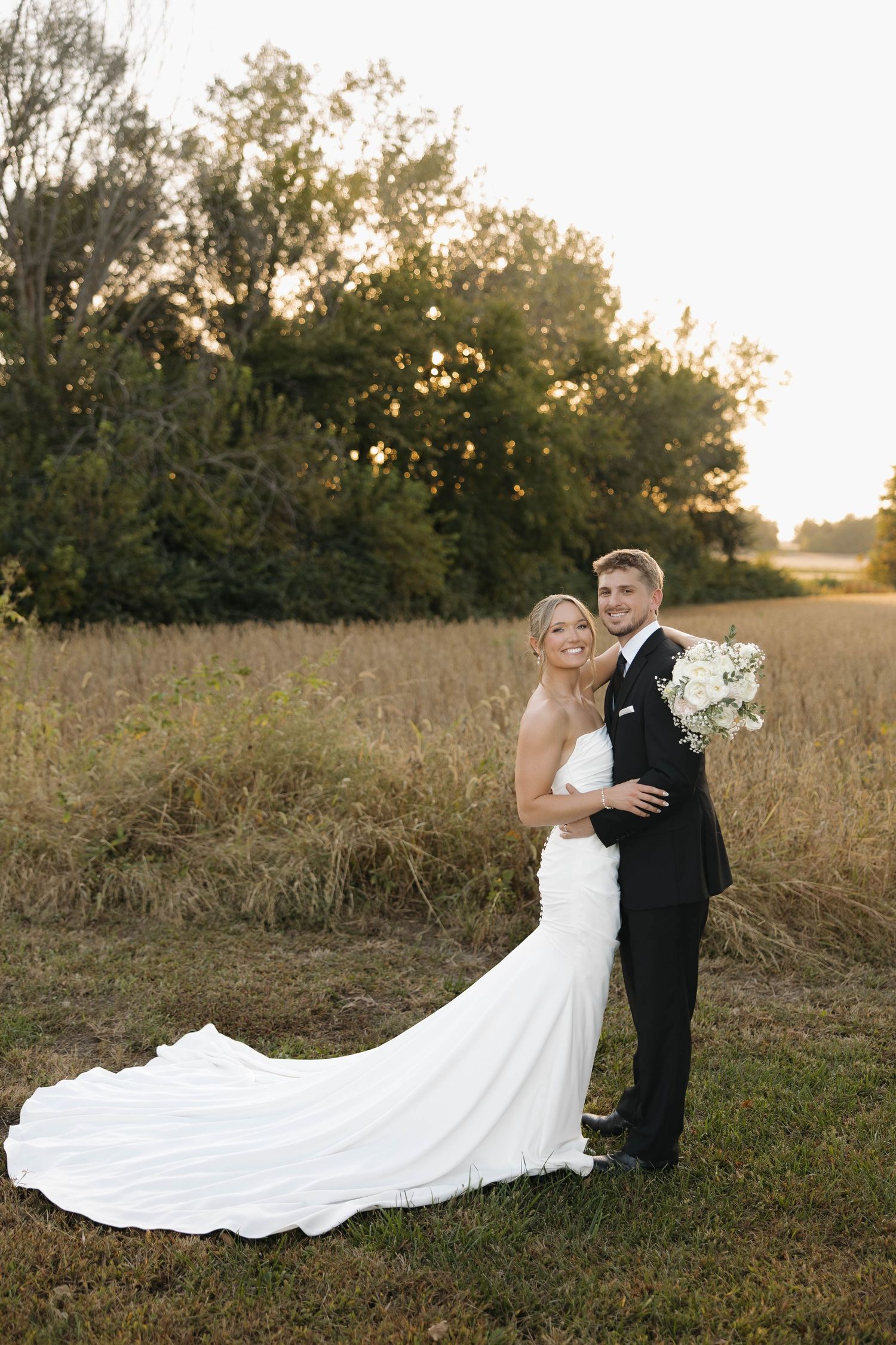 A bride and groom embrace in a golden field at sunset, with her flowing white dress train creating an elegant silhouette.