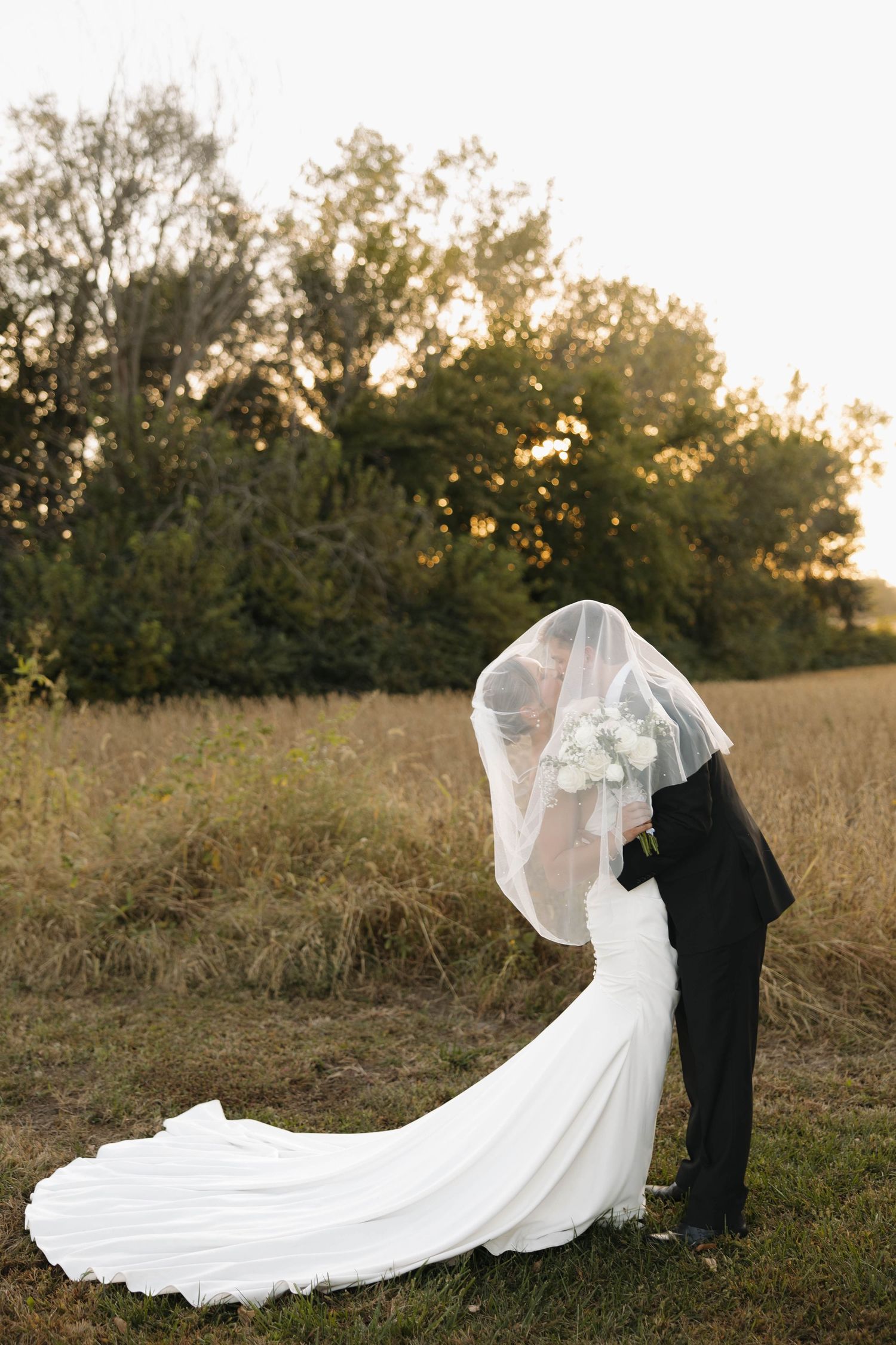 Wedding couple poses in a golden field at sunset with bride's veil flowing in the breeze.