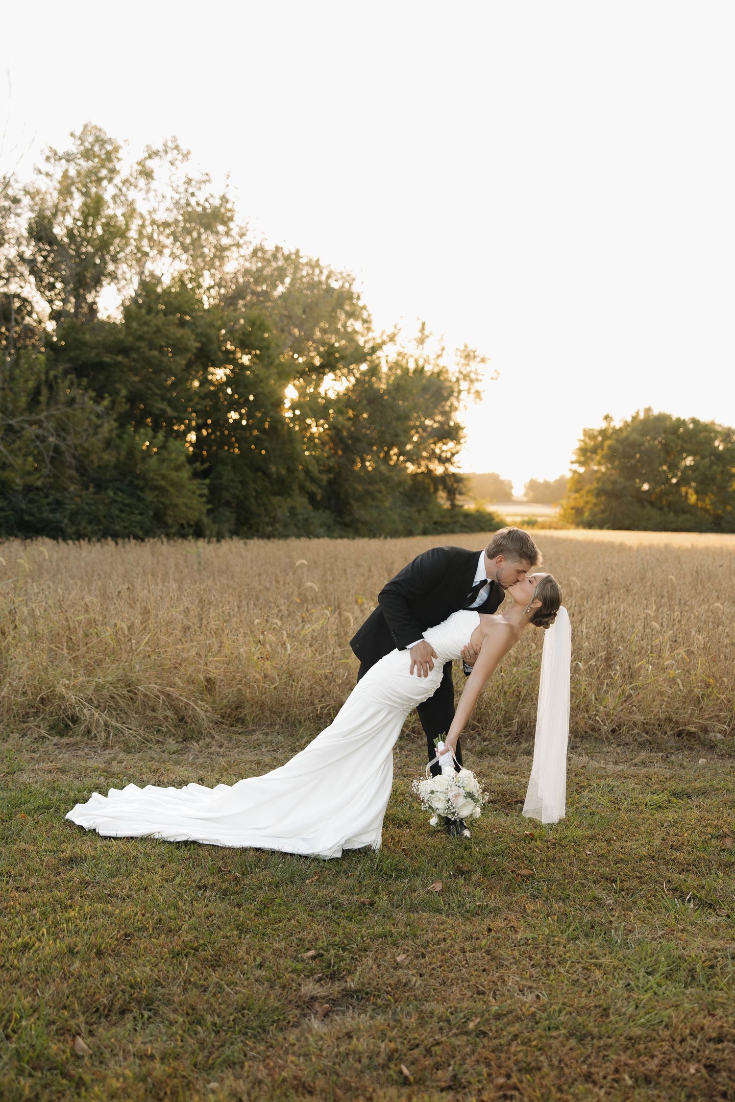 Newlyweds share a romantic dip kiss in a golden field at sunset.