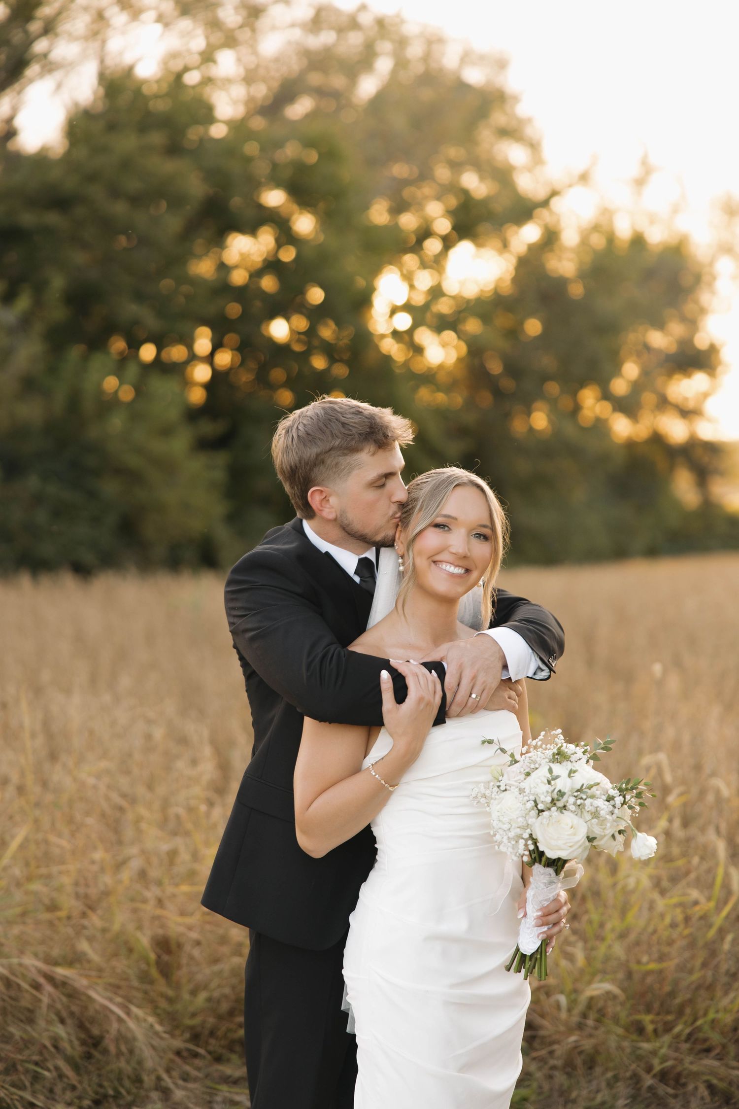 A tender moment captured between newlyweds in a sunlit field with white flowers and formal attire.